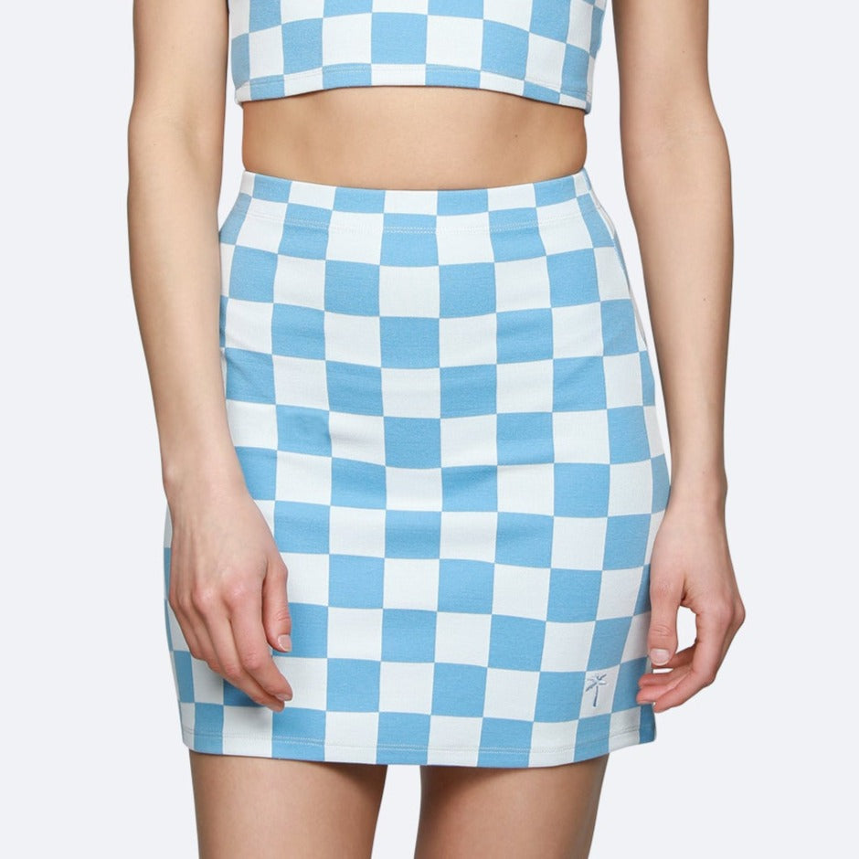 Eine Person, die ein passendes blau-weiß kariertes Crop-Top und einen Minirock trägt, steht mit entspannten Armen an der Seite vor einem schlichten Hintergrund.