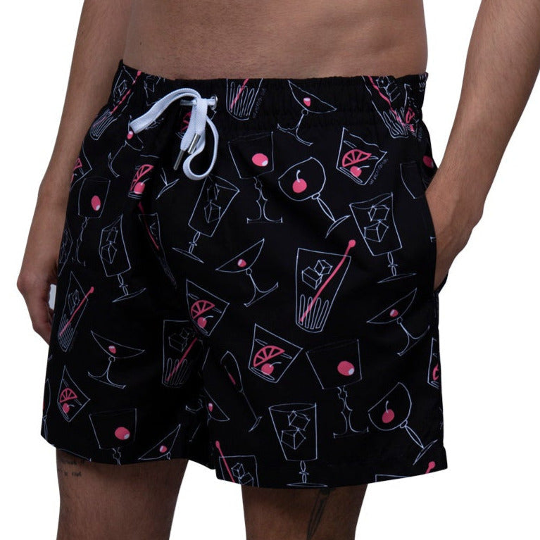 Eine Person, die eine schwarze Badehose mit weißem Kordelzug trägt, auf der ein buntes Muster von Cocktails abgebildet ist, darunter Martini- und Old Fashioned-Gläser mit Kirschen und Zitrusscheiben.