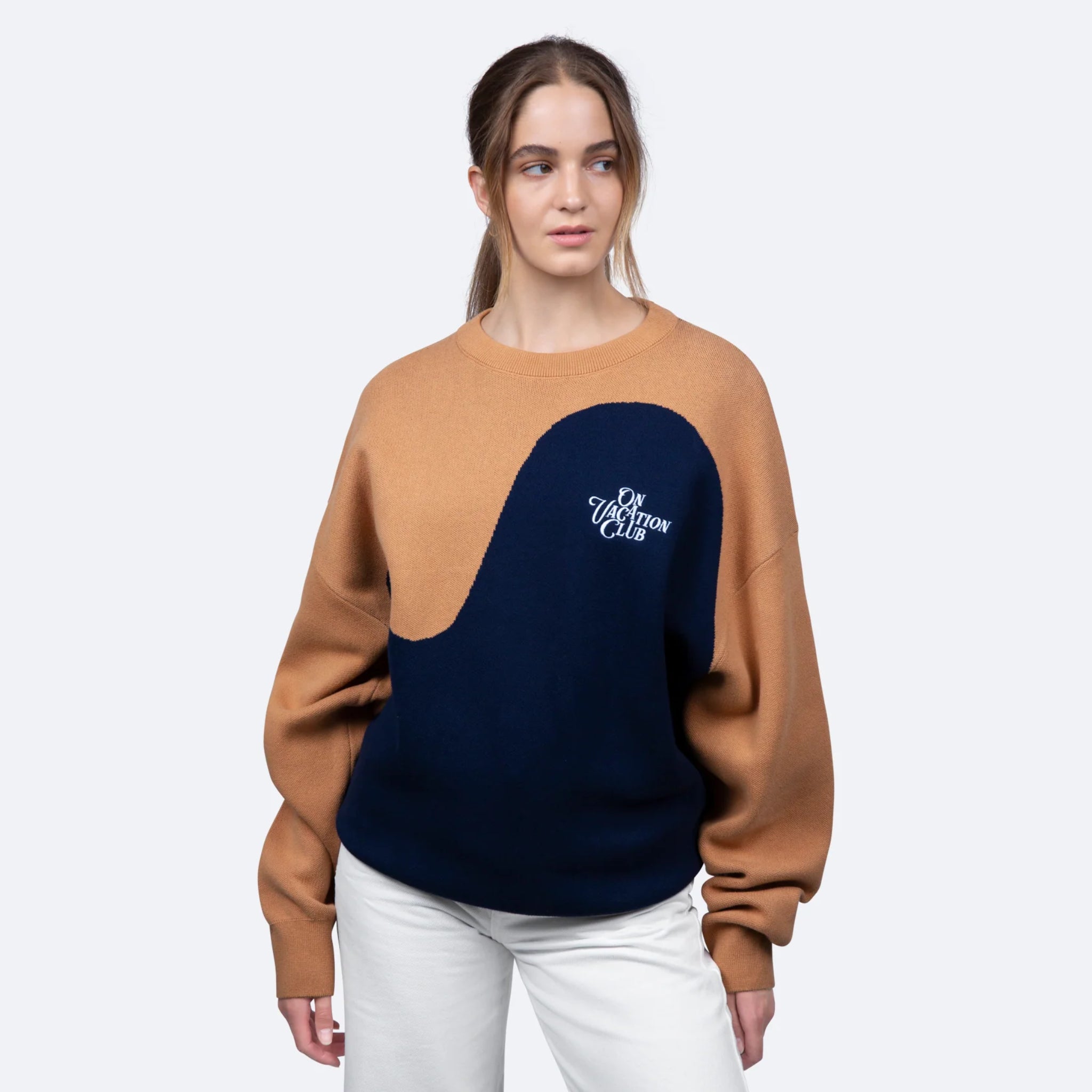 Frau trägt einen Pullover mit braun-blauem Design und Aufschrift "On Vacation Club", dazu weiße Hose.