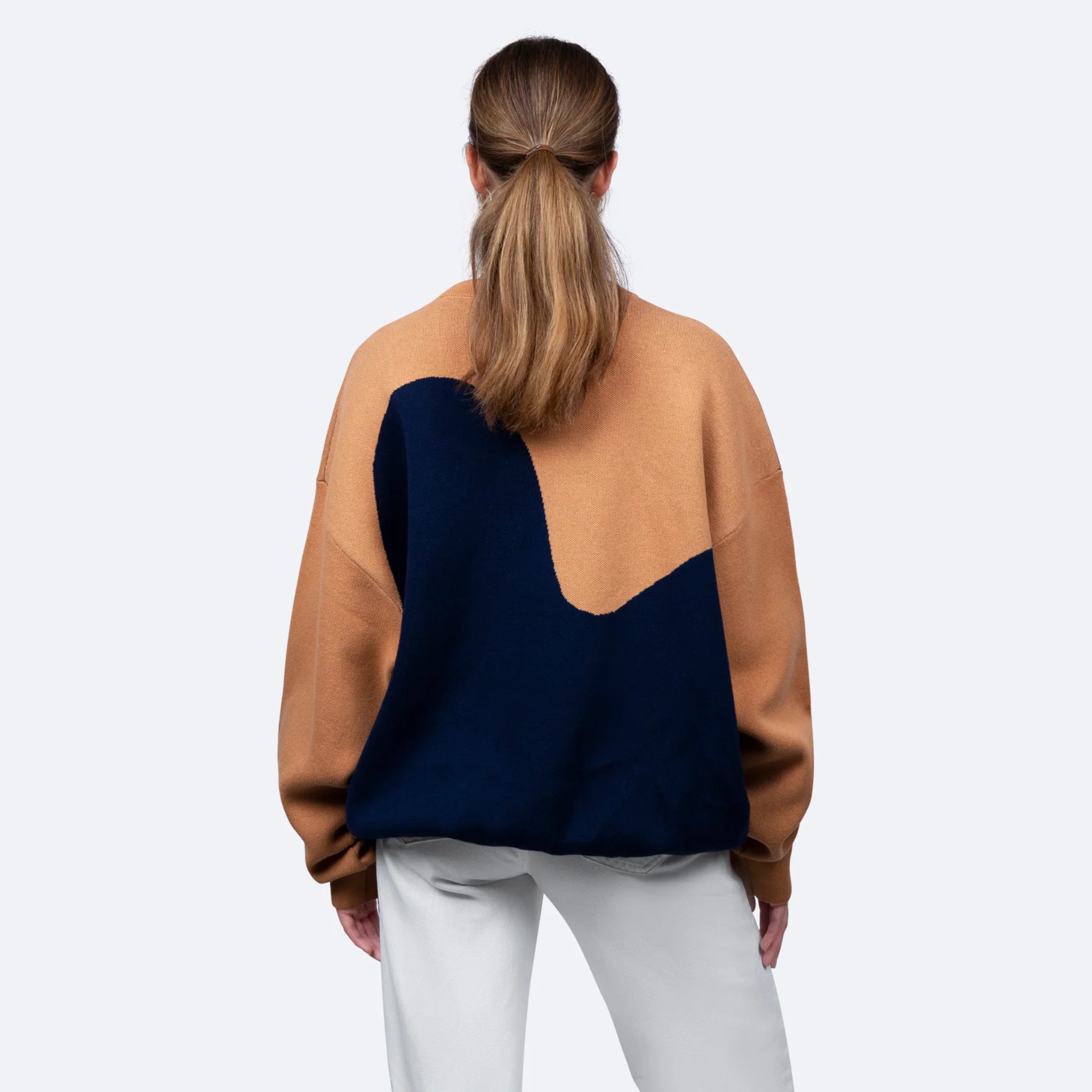 Person von hinten in einem braunen und dunkelblauen Pullover mit weißer Hose, Haare im Pferdeschwanz gebunden.