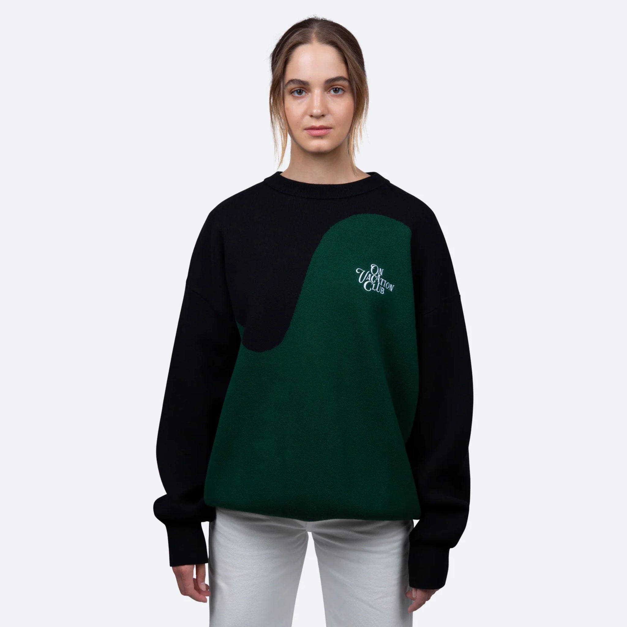 Frau trägt einen schwarz-grünen Pullover mit Aufschrift "On Vacation Club" und weiße Hose vor neutralem Hintergrund.