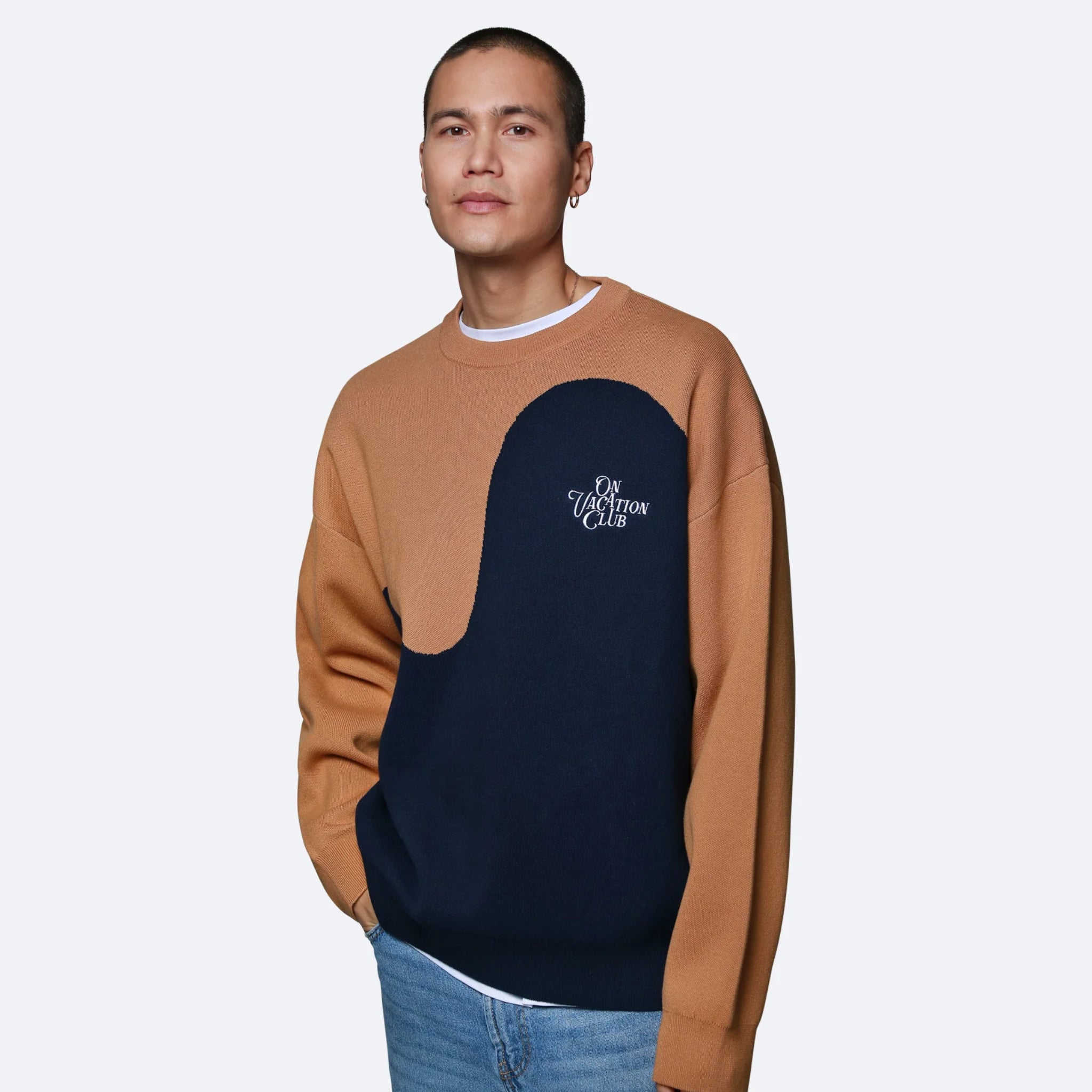 Ein Mann trägt einen braun-blauen Pullover mit Schriftzug "Vacation Club" und blaue Jeans vor einem hellgrauen Hintergrund.