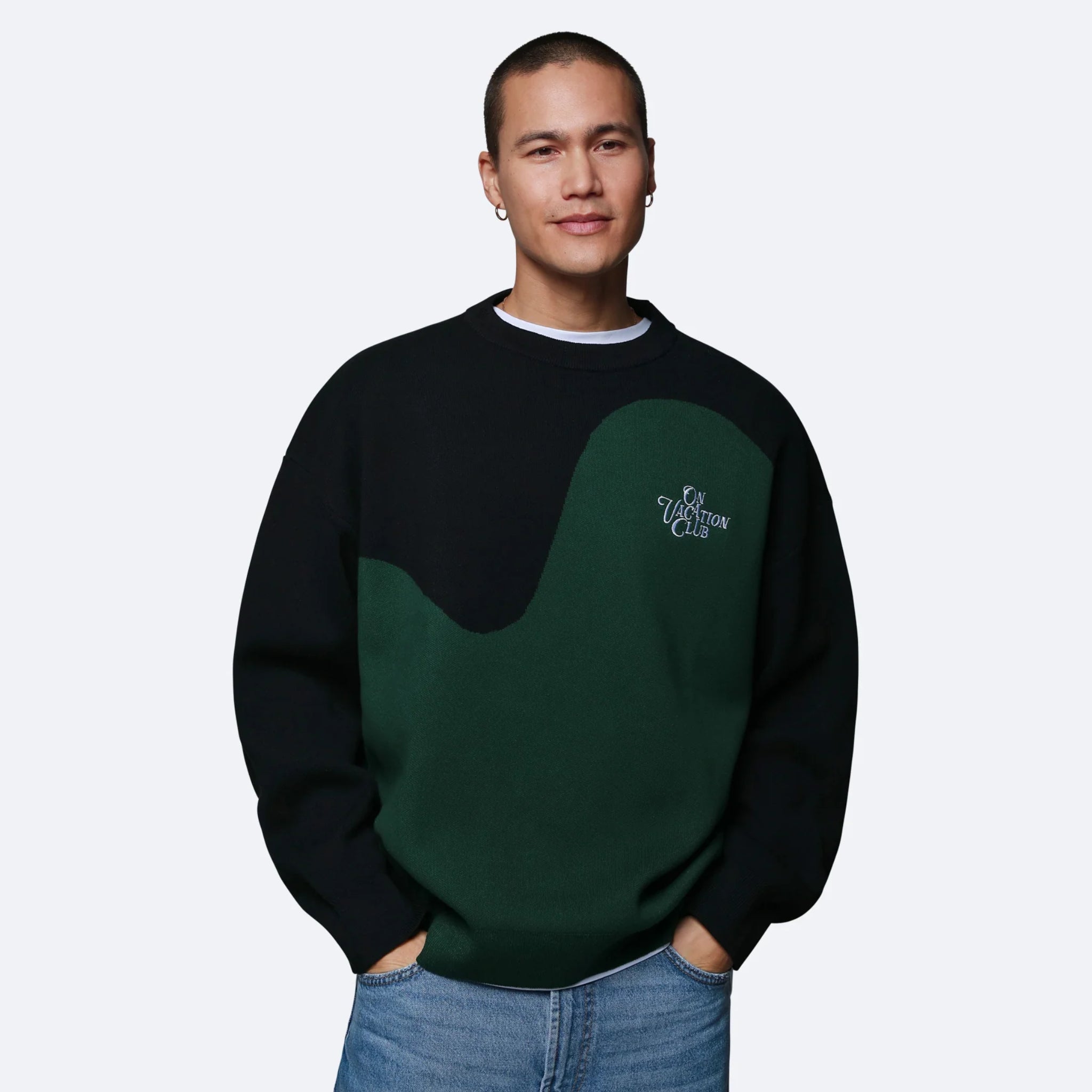 Person in a zweifarbigem Sweatshirt mit der Aufschrift „On Vacation Club“ und blauer Jeans vor einem grauen Hintergrund.