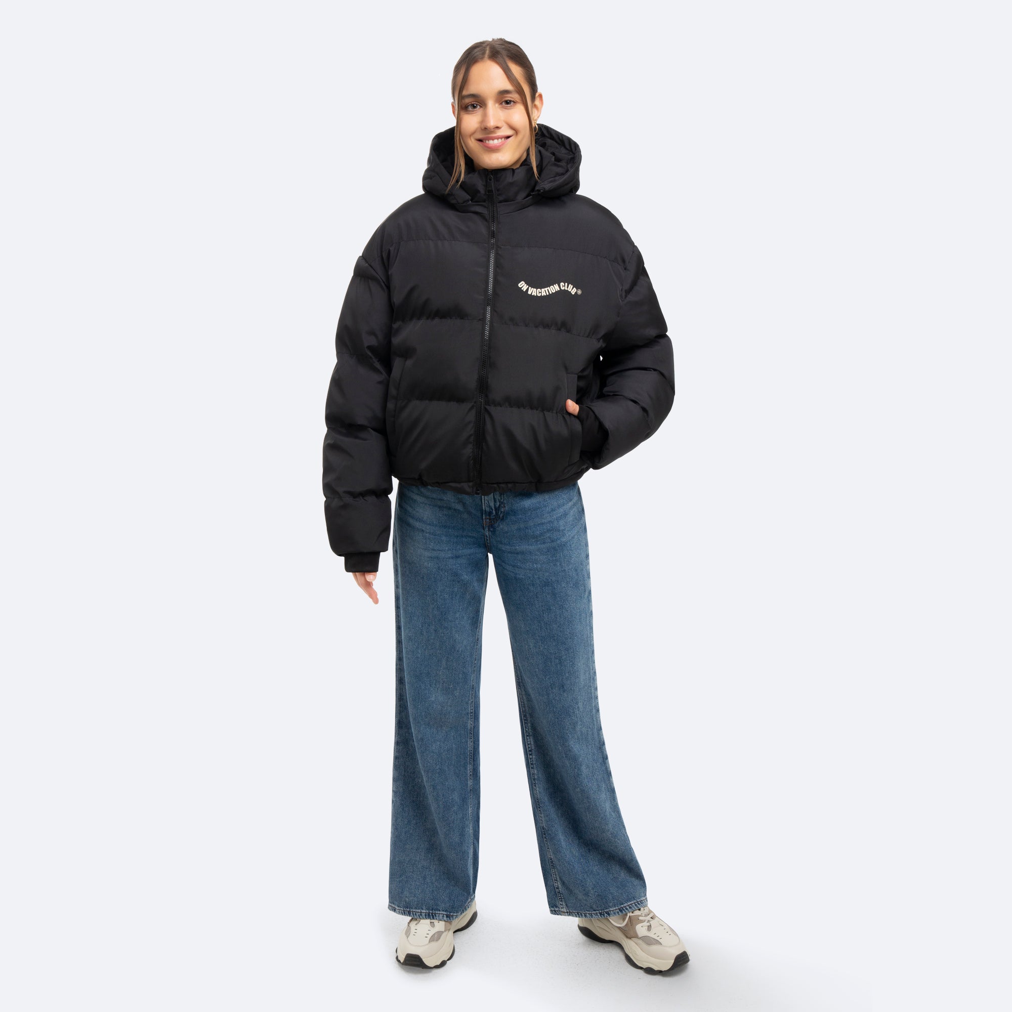 Eine Person, die eine schwarze Pufferjacke mit Kapuze, blaue Jeans mit weitem Bein und helle Turnschuhe trägt, steht lächelnd vor einem schlichten hellen Hintergrund.