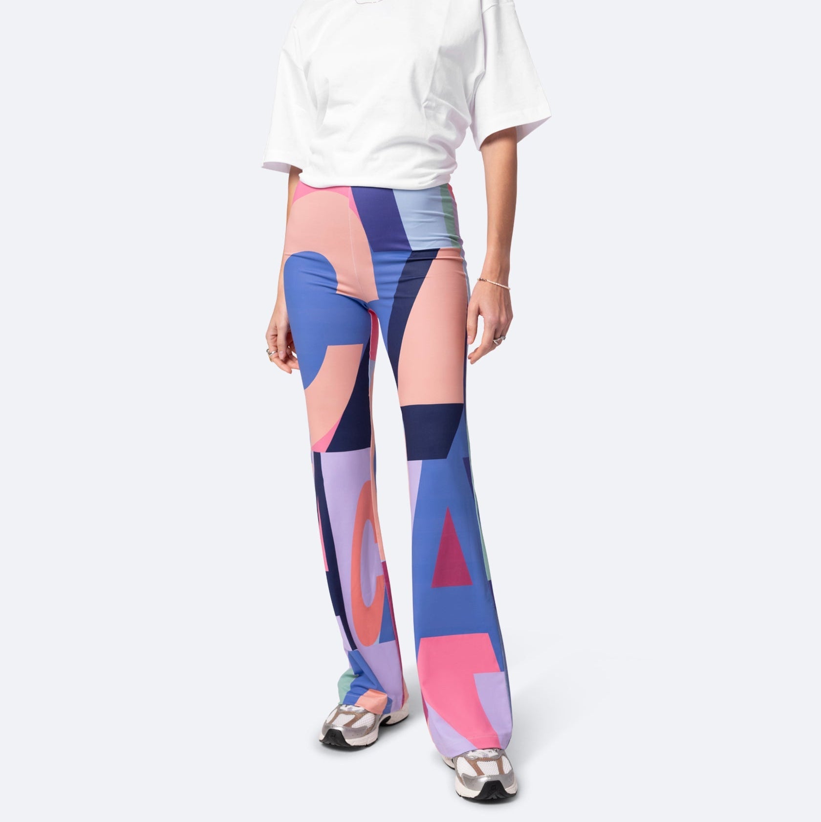 Eine Person, die ein lockeres weißes T-Shirt, eine bunte, geometrisch gemusterte Schlaghose in Blau-, Rosa- und Lila-Tönen und weiße Turnschuhe trägt, steht vor einem schlichten hellen Hintergrund.