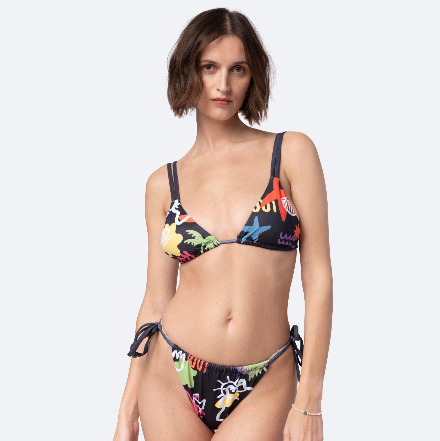 Eine Frau mit kurzen braunen Haaren trägt einen farbenfrohen Bikini mit Cartoon-Aufdruck und seitlichen Bändern. Sie steht selbstbewusst vor einem schlichten, hellen Hintergrund und blickt direkt in die Kamera.