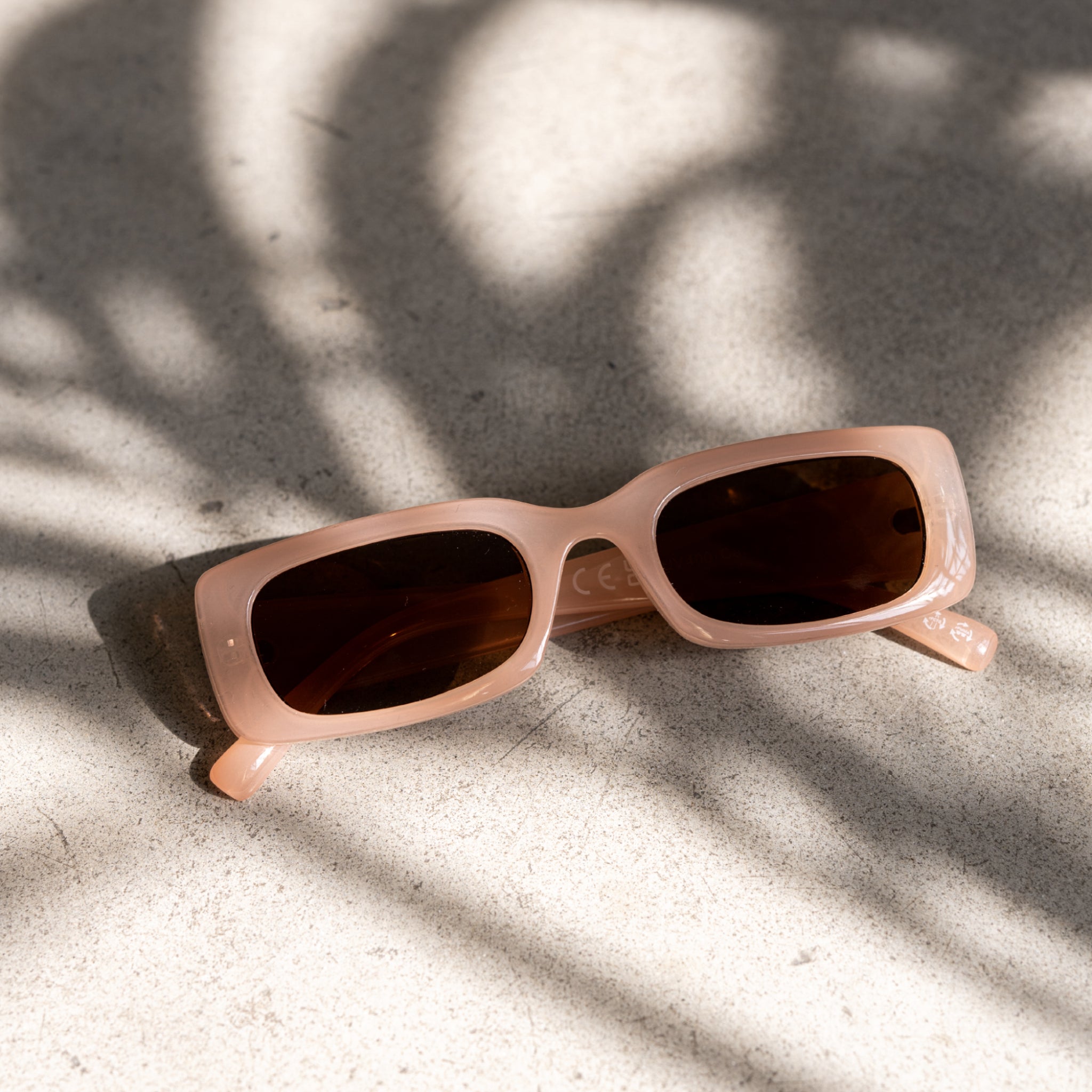 Eine beige Sonnenbrille liegt auf einem hellen, strukturierten Untergrund im Schatten von Pflanzen.