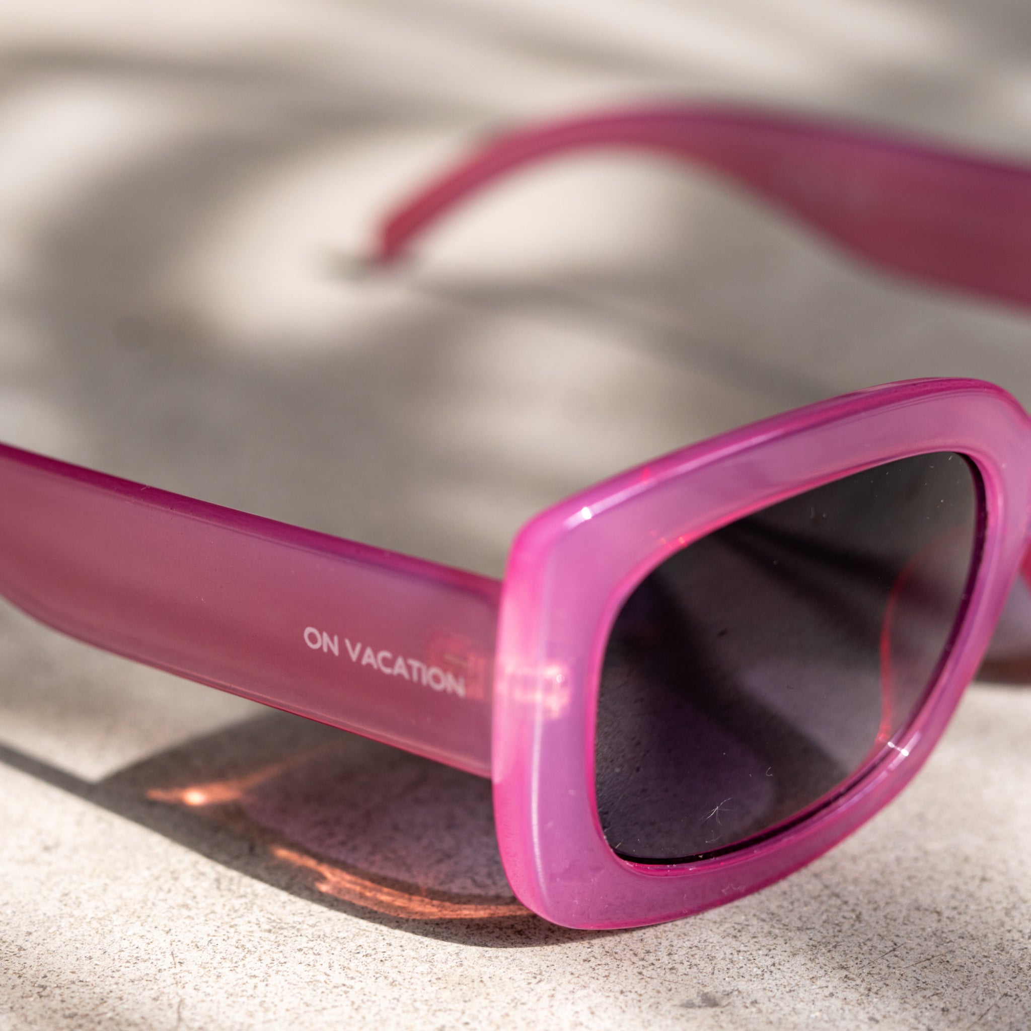 Pinkfarbene Sonnenbrille mit der Aufschrift "ON VACATION" auf einem beigen Untergrund.