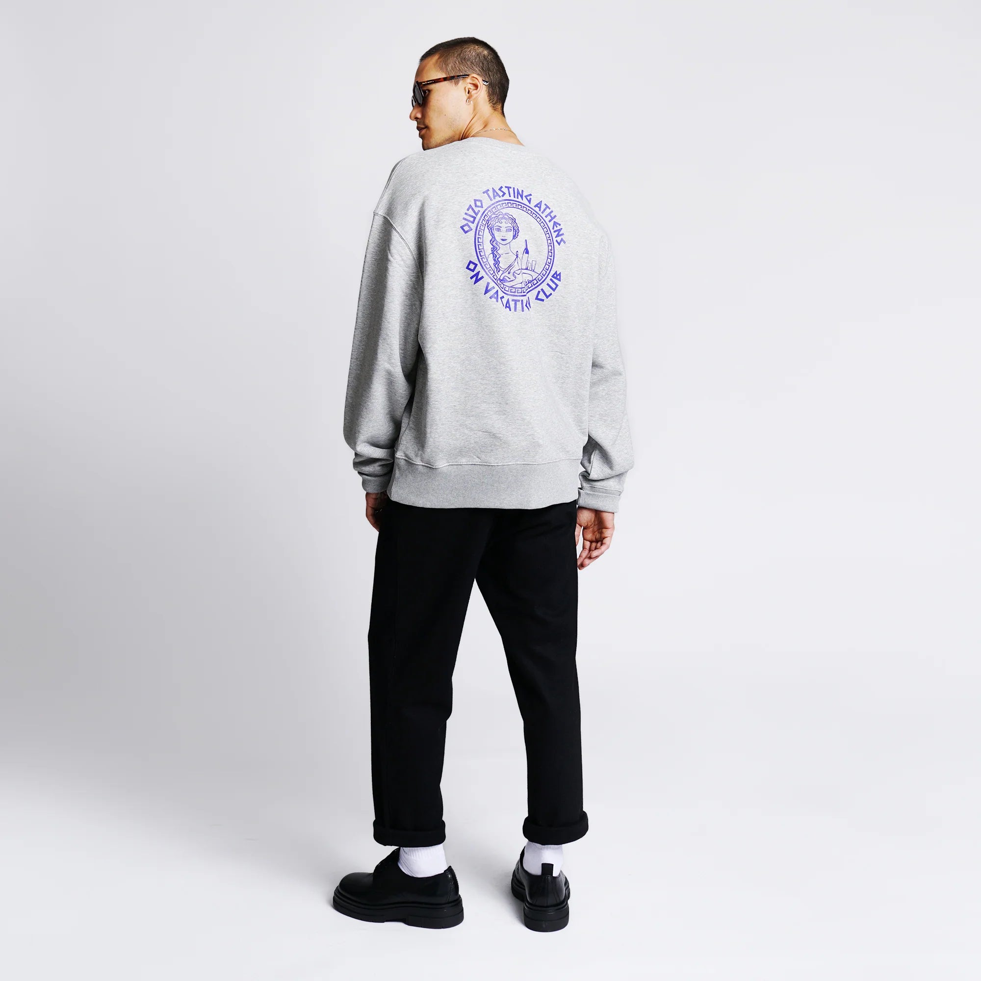 Eine Person in grauem Sweatshirt mit blauem Aufdruck auf dem Rücken, schwarzer Hose und schwarzen Schuhen.