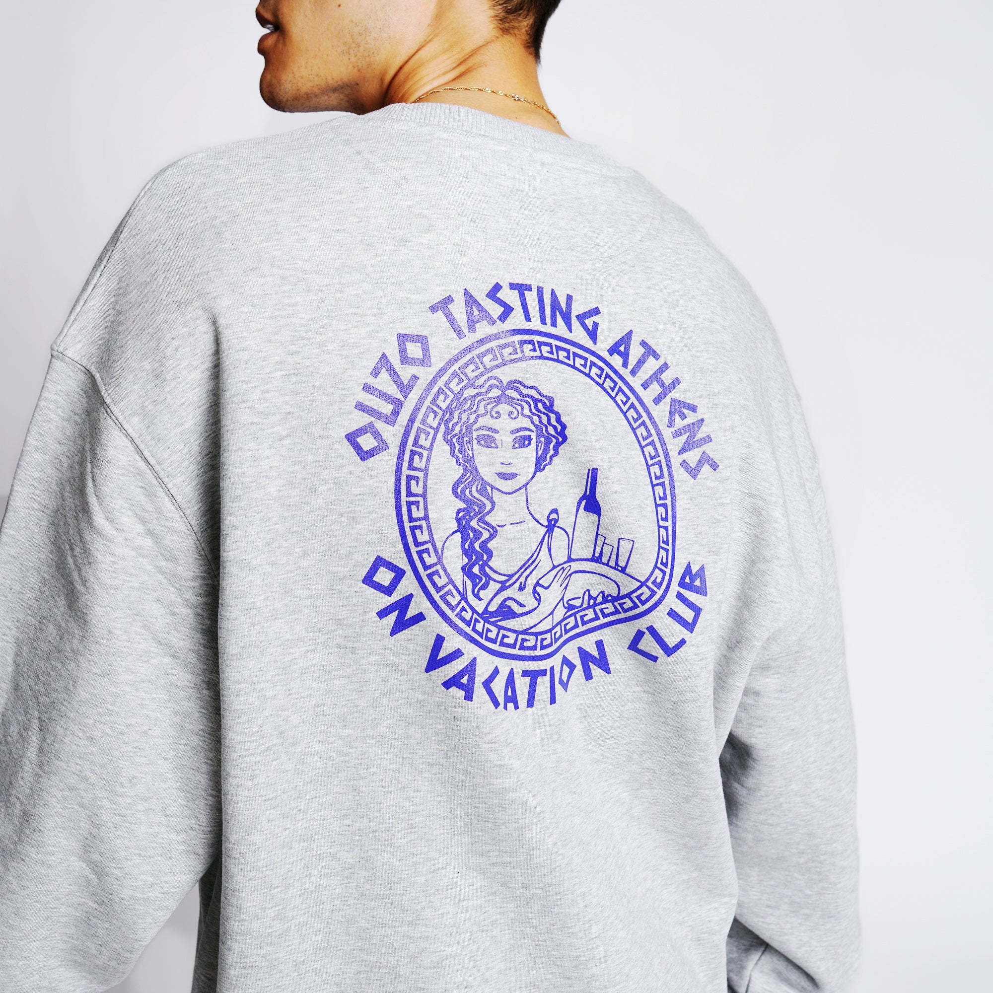 Rückseite eines grauen Pullovers mit lila Grafik einer Frau und dem Text "Ouzo Tasting Athens On Vacation Club".