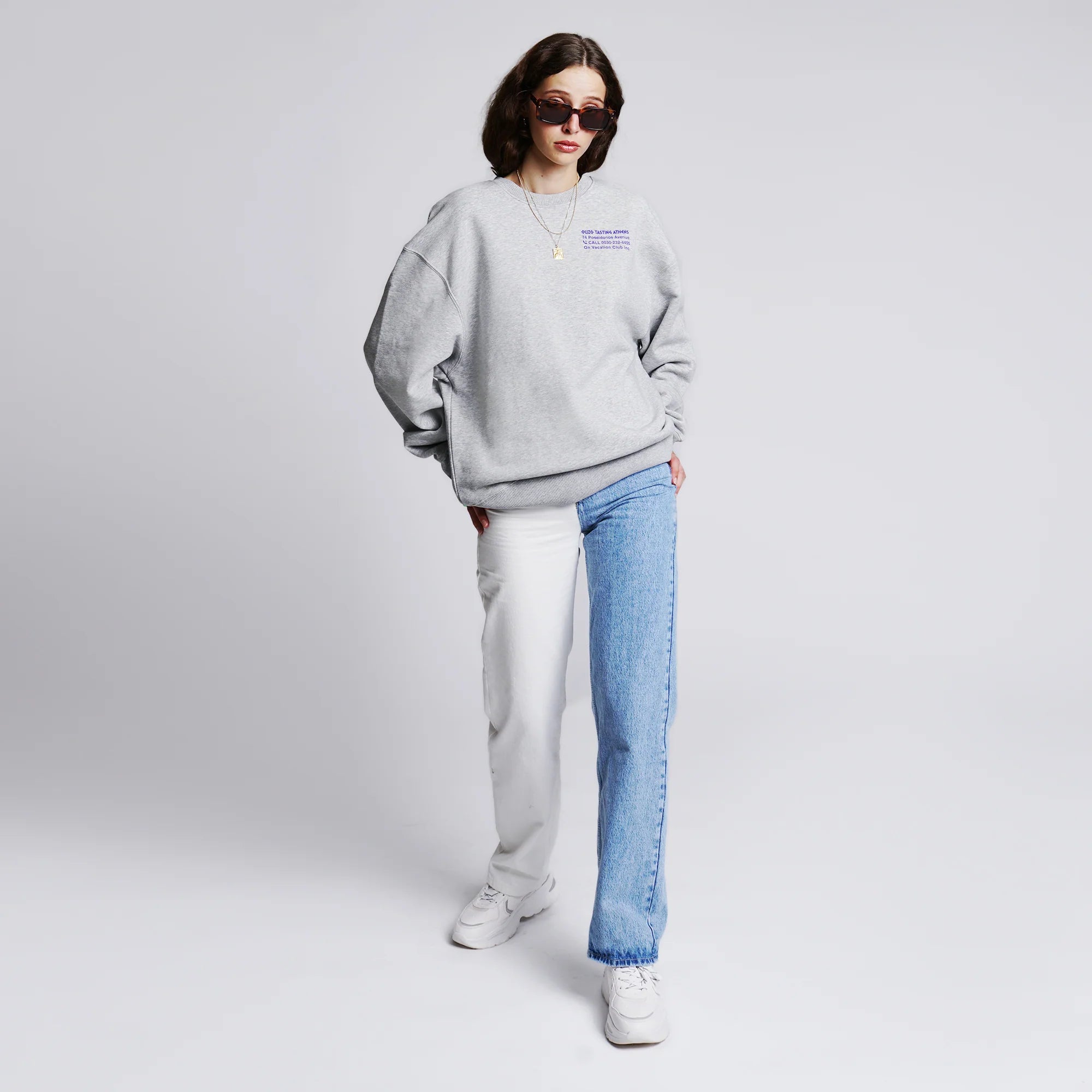 Person in oversized grauem Sweatshirt, geteilter Jeans und weißen Sneakern, mit Sonnenbrille und neutralem Hintergrund.