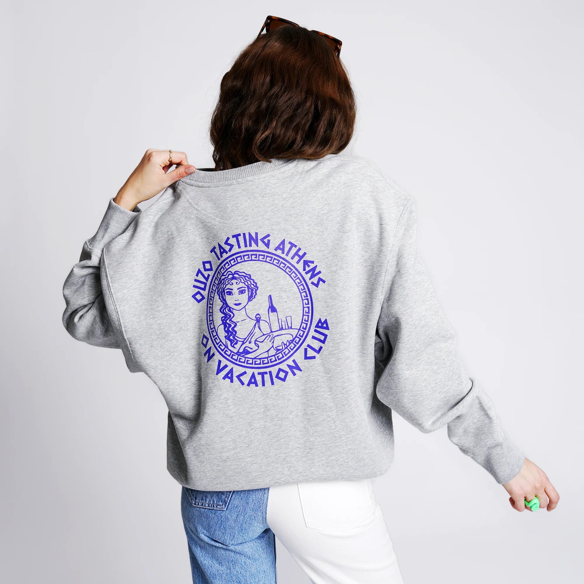 Eine Frau trägt einen grauen Pullover mit violettem Aufdruck "OUZO TASTING ATHENS ON VACATION CLUB" auf dem Rücken.