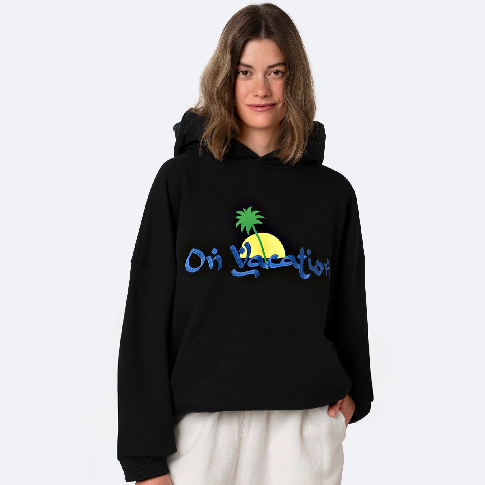 Eine Frau mit hellbraunem Haar trägt einen schwarzen Kapuzenpulli mit einer gelben Sonne, einer grünen Palme und der Aufschrift "On Vacation" in blauer Schrift. Sie steht vor einem schlichten hellen Hintergrund und lächelt leicht.