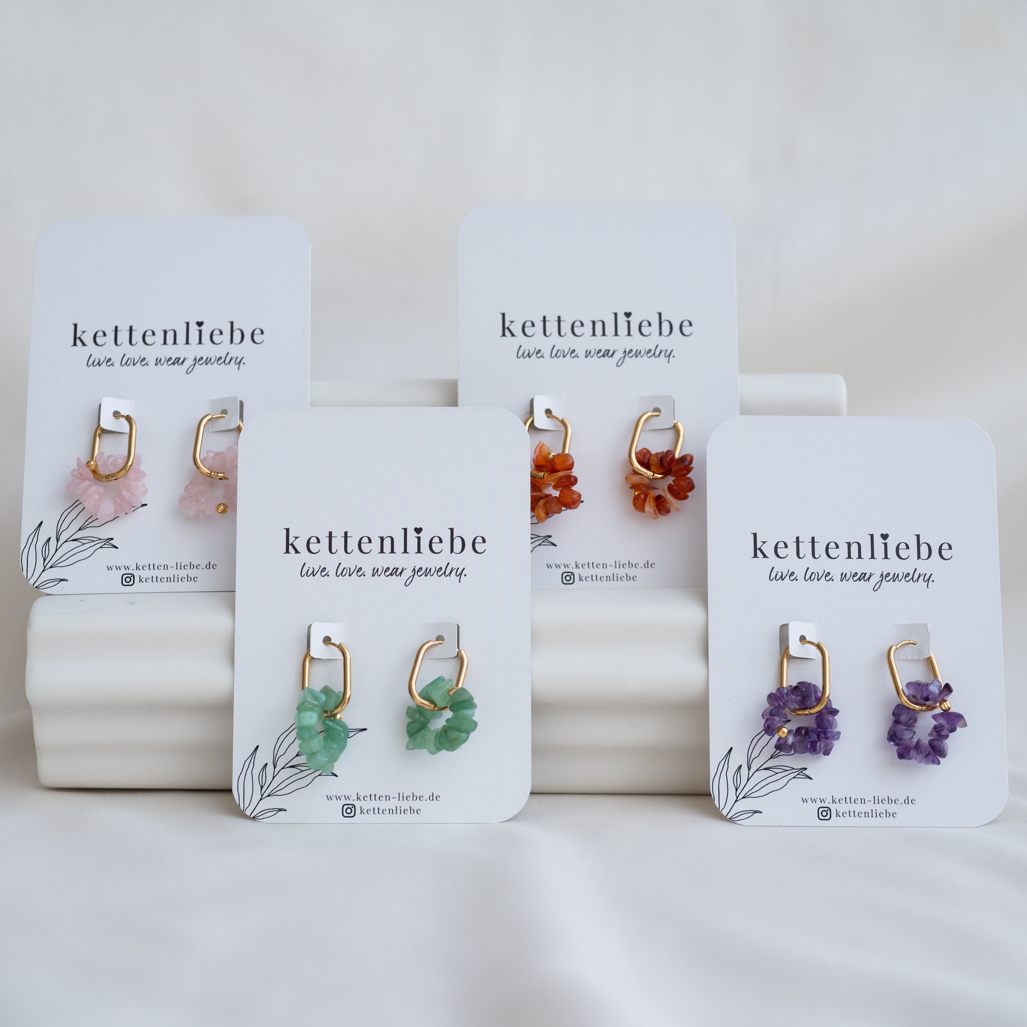 Vier Paare farbenfroher Edelstein-Ohrringe auf Display-Karten mit der Aufschrift "kettenliebe", jeweils mit gebündelten Steinen in Rosa, Orange, Grün und Lila. Die Ohrringe sind auf weißen Plattformen angeordnet.