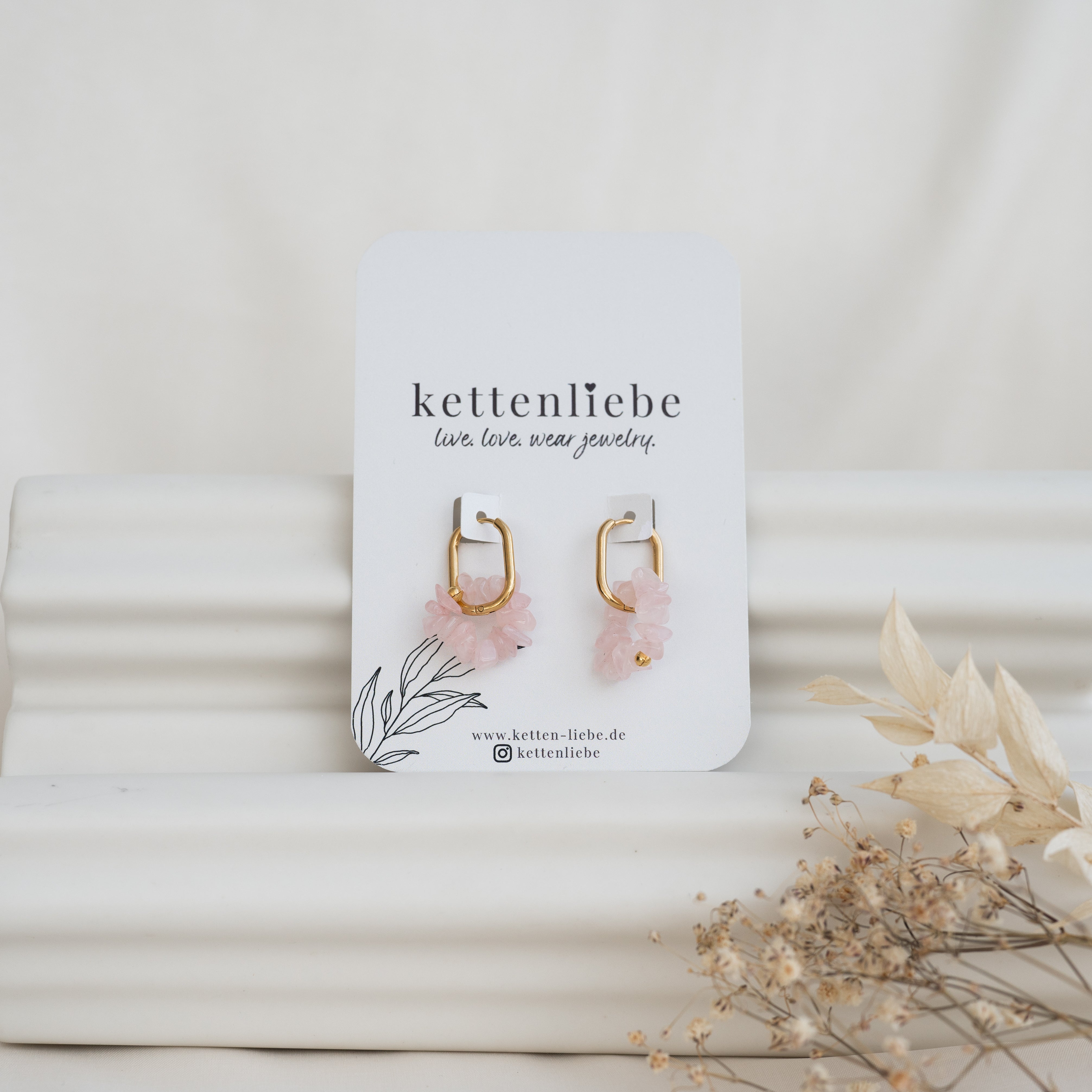 Ein Paar goldene Ohrringe mit rosafarbenen Blumenanhängern ist auf einer weißen Karte mit der Aufschrift kettenliebe abgebildet, die vor einem weißen, strukturierten Hintergrund mit getrockneten Blumen im Vordergrund steht.