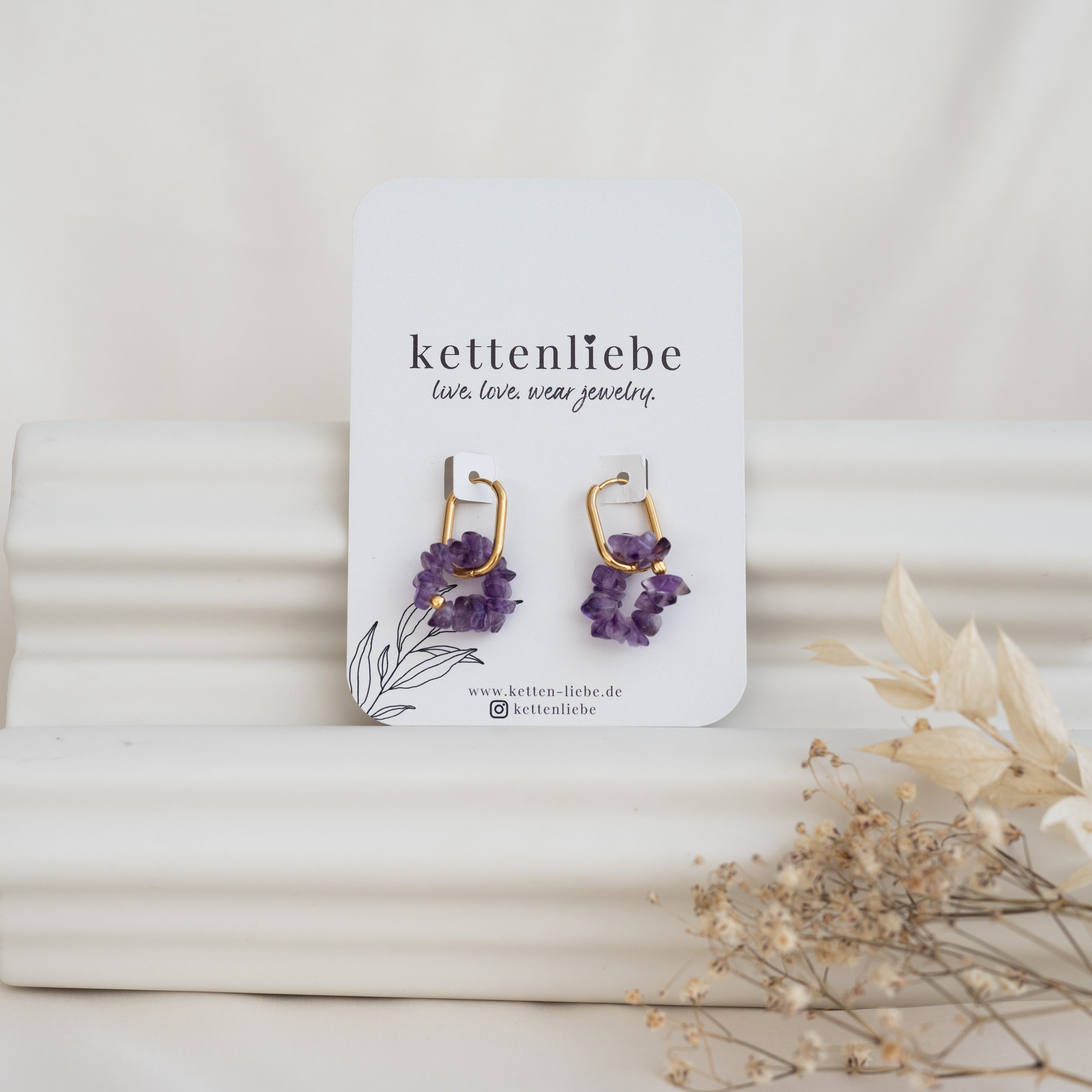 Ein Paar goldene und violette Edelsteinohrringe auf einer Karte mit der Aufschrift "kettenliebe" und "live. love. wear jewelry" vor einem weißen Hintergrund mit Trockenblumen im Vordergrund.