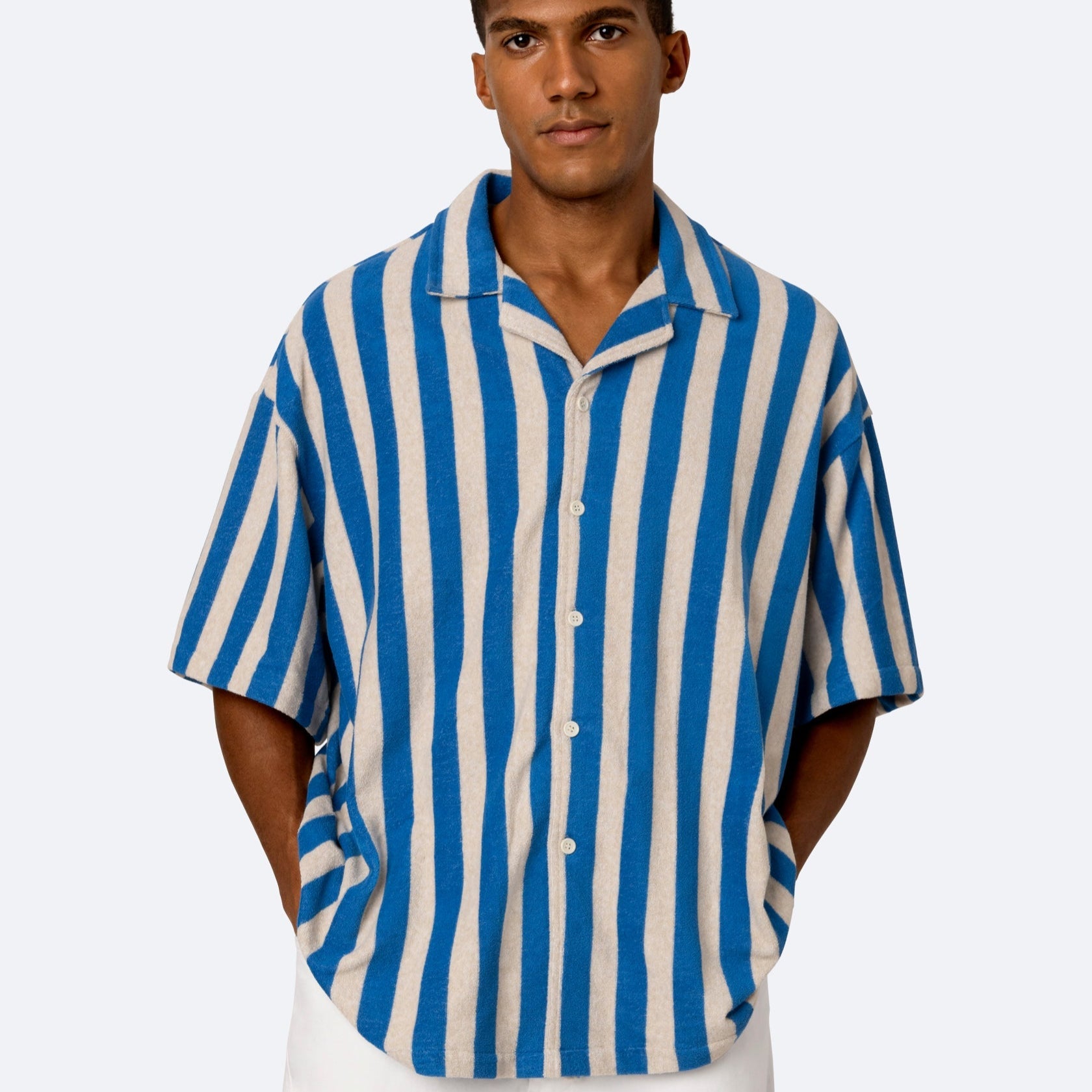 Ein Mann steht mit den Händen in den Taschen, trägt ein blau-beigefarbenes, vertikal gestreiftes, kurzärmeliges Button-up-Hemd und eine weiße Hose vor einem schlichten, hellen Hintergrund.