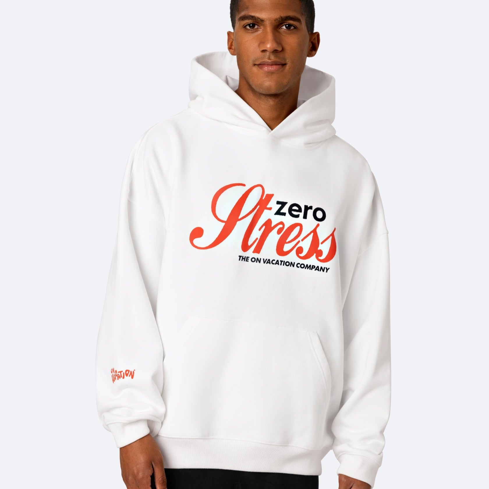 Ein Mann, der einen weißen Kapuzenpulli mit der Aufschrift "Zero Stress THE ON VACATION COMPANY" in großen roten und schwarzen Buchstaben auf der Vorderseite trägt, steht vor einem schlichten hellgrauen Hintergrund.