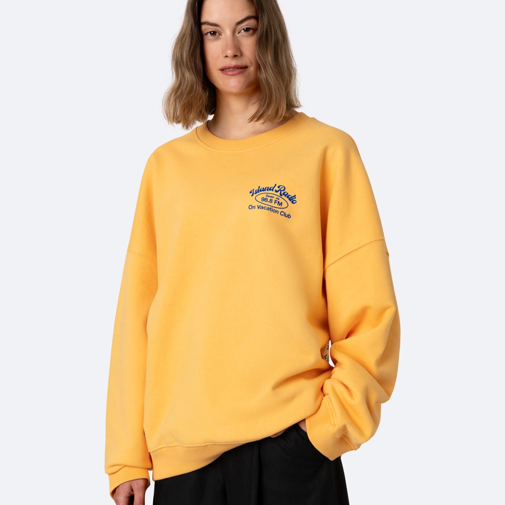 Eine Frau mit schulterlangen braunen Haaren trägt ein locker sitzendes gelbes Sweatshirt mit blauer Aufschrift auf der linken Brust und eine schwarze Hose vor einem schlichten hellen Hintergrund.