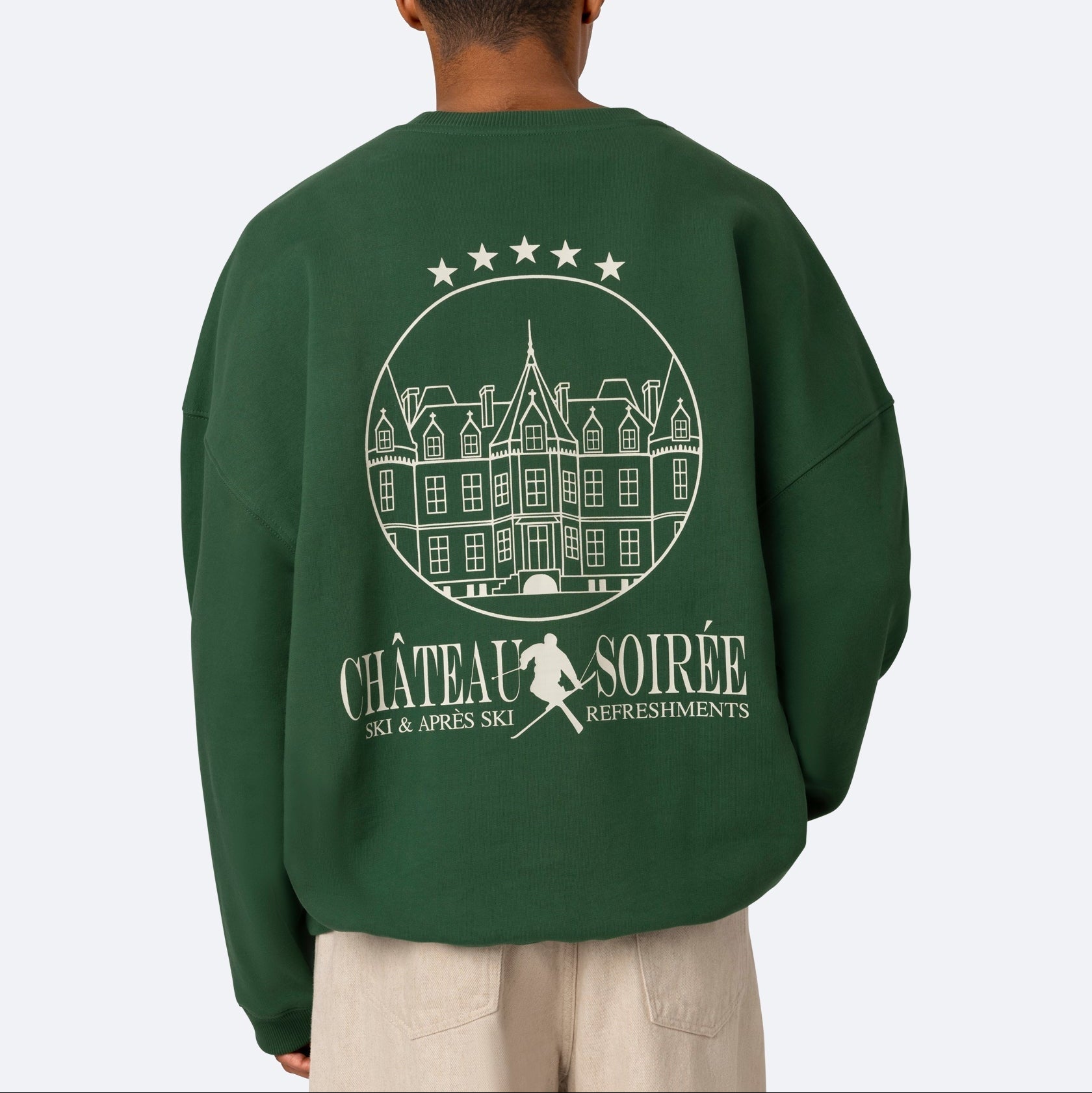 Eine Person trägt ein grünes Sweatshirt mit einer weißen Grafik eines Schlosses, fünf Sternen und dem Text "CHÂTEAU SOIRÉE SKI & APRÈS SKI REFRESHMENTS" auf dem Rücken.