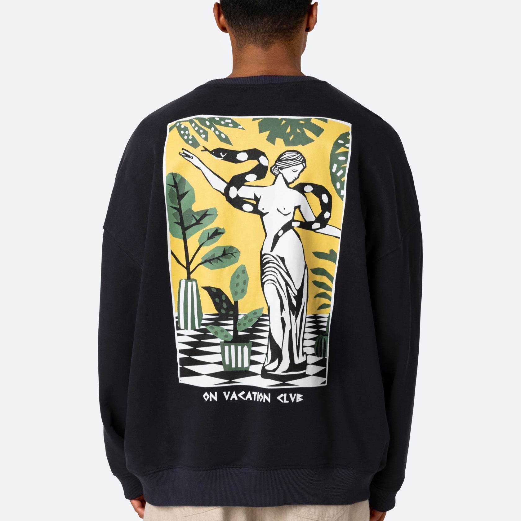 Eine Person trägt ein schwarzes Sweatshirt mit einer großen Grafik auf dem Rücken, die eine stilisierte Statue, Pflanzen und einen Schachbrettboden zeigt, zusammen mit dem Text "ON VACATION CLUB" am unteren Rand.
