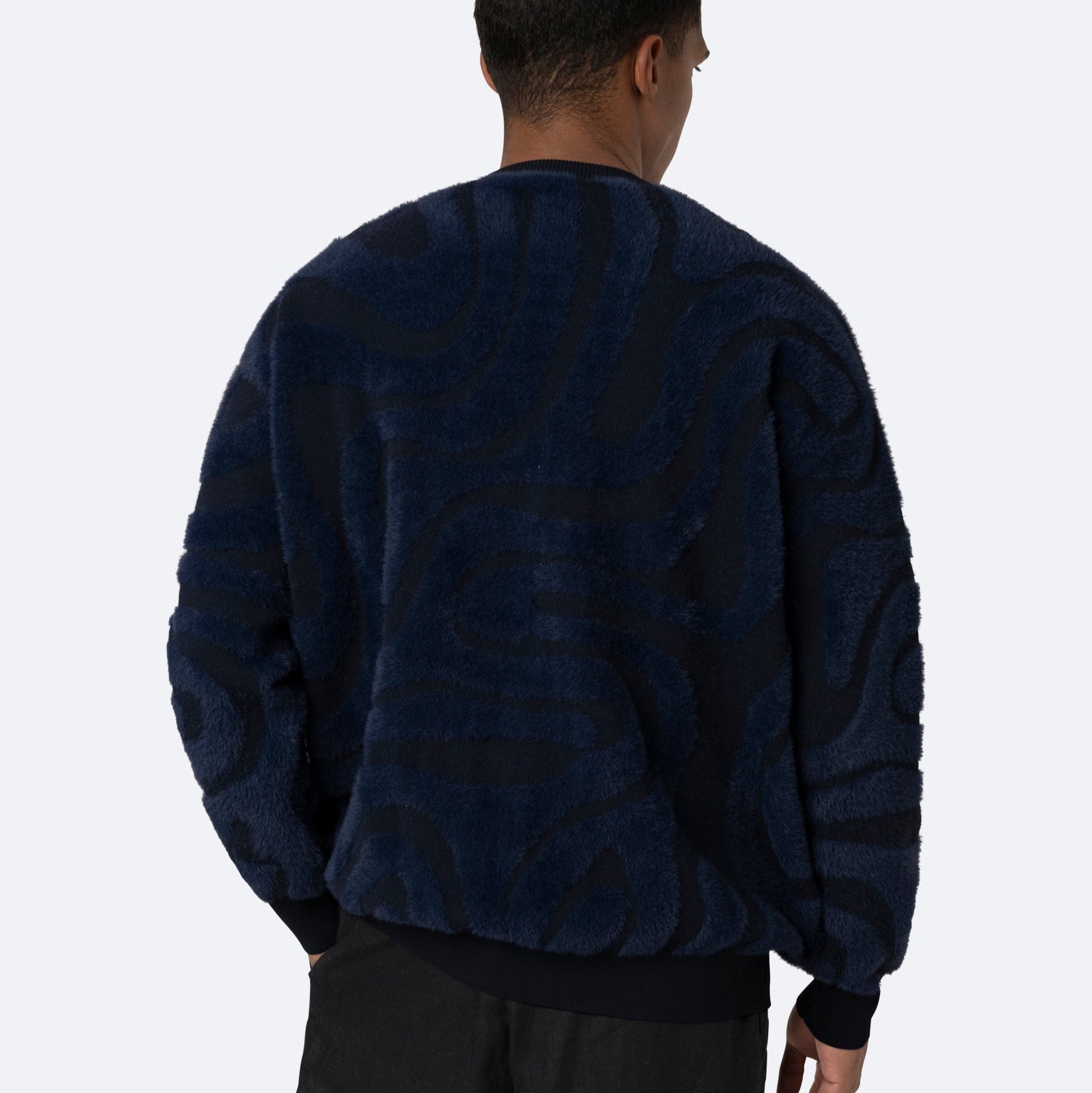 Eine Person, die ein dunkles, wuscheliges Sweatshirt mit wellenförmigen, abstrakten Mustern in marineblauen Farbtönen trägt, steht mit dem Gesicht zur Seite und zeigt die Rückseite des Kleidungsstücks vor einem schlichten hellen Hintergrund.