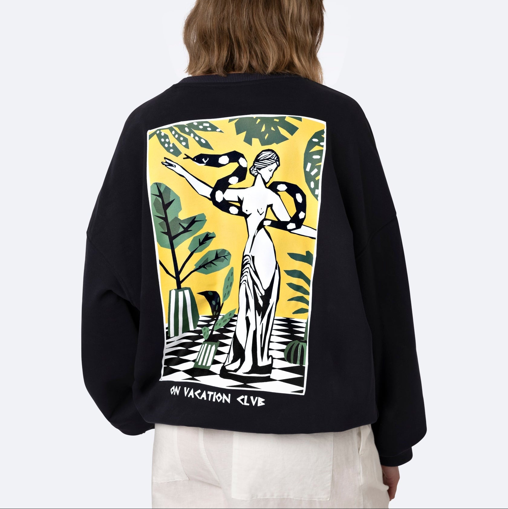 Eine Person mit schulterlangen braunen Haaren trägt ein schwarzes Sweatshirt mit einer farbenfrohen Grafik einer griechischen Statue mit einer Schlange, umgeben von tropischen Pflanzen, und dem Text ON VACATION CLUB am unteren Rand.