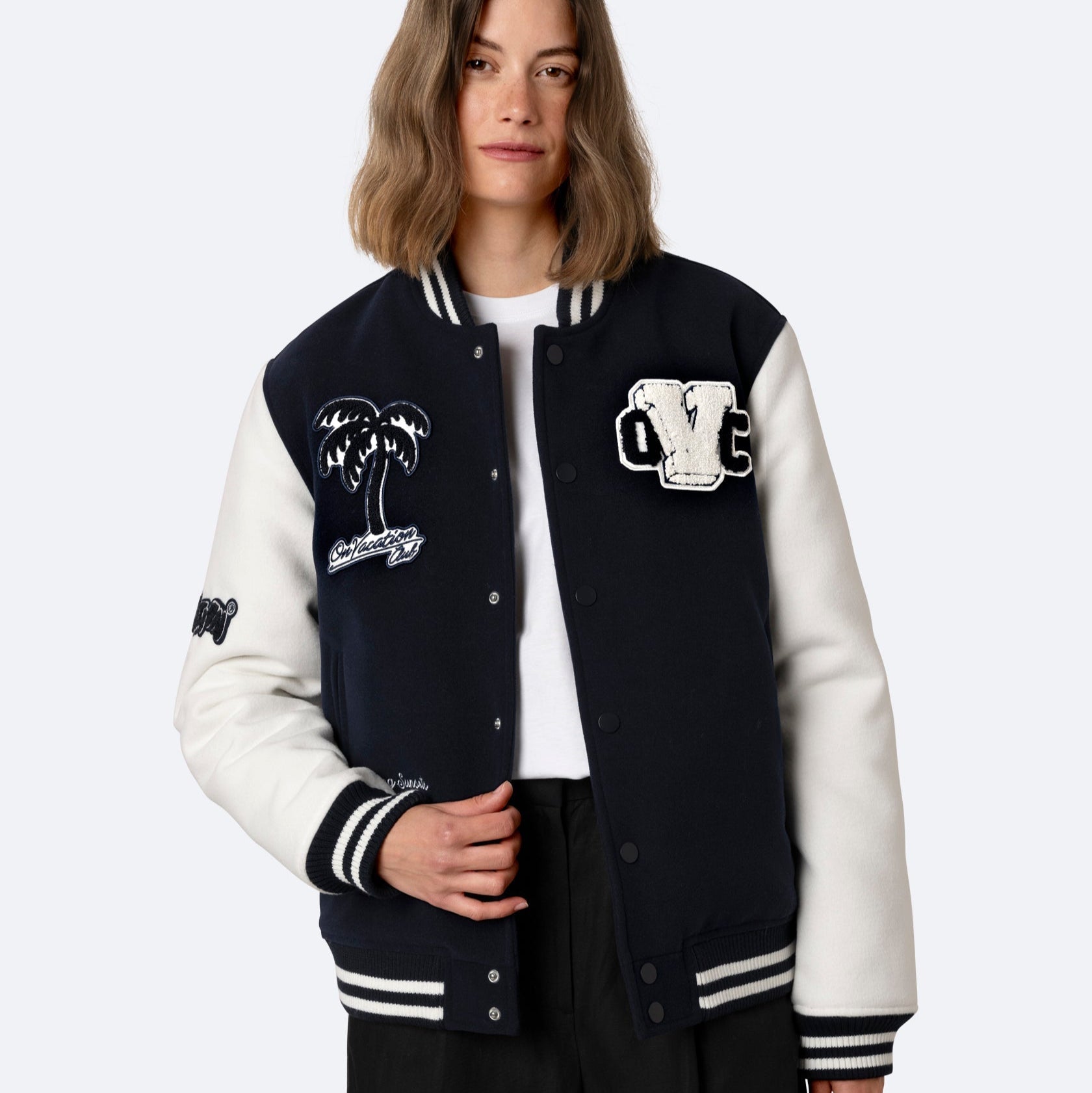 Eine Frau mit schulterlangem Haar trägt eine marineblaue und weiße Sportjacke mit aufgestickten Aufnähern, darunter eine Palme und Buchstaben, über einem weißen Hemd und einer schwarzen Hose, die vor einem unifarbenen Hintergrund stehen.