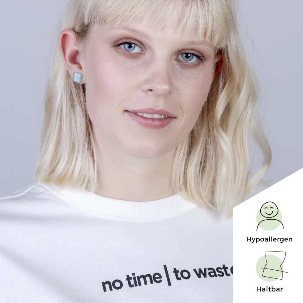 Eine Frau mit blonden Haaren trägt blaue, quadratische Ohrringe und ein weißes Hemd mit der Aufschrift "No time | to waste". In der unteren rechten Ecke sind Symbole für "hypoallergen" und "langlebig" zu sehen.