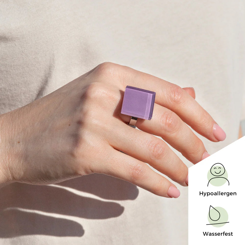 Eine Hand, die einen großen, quadratischen, violetten Ring trägt, wird vor einem hellen Hintergrund gezeigt. In der Ecke zeigen Symbole an, dass der Ring hypoallergen und wasserfest ist, mit den Worten Hypoallergen und Wasserfest.