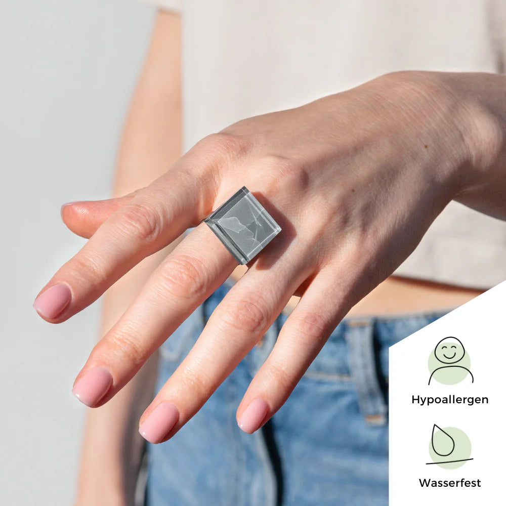 Eine Hand mit hellrosa lackierten Nägeln trägt einen großen, quadratischen, grauen Steinring. Der Hintergrund ist hell und zeigt eine Person am Handgelenk und eine blaue Jeans. Symbole zeigen an, dass der Ring hypoallergen und wasserdicht ist.