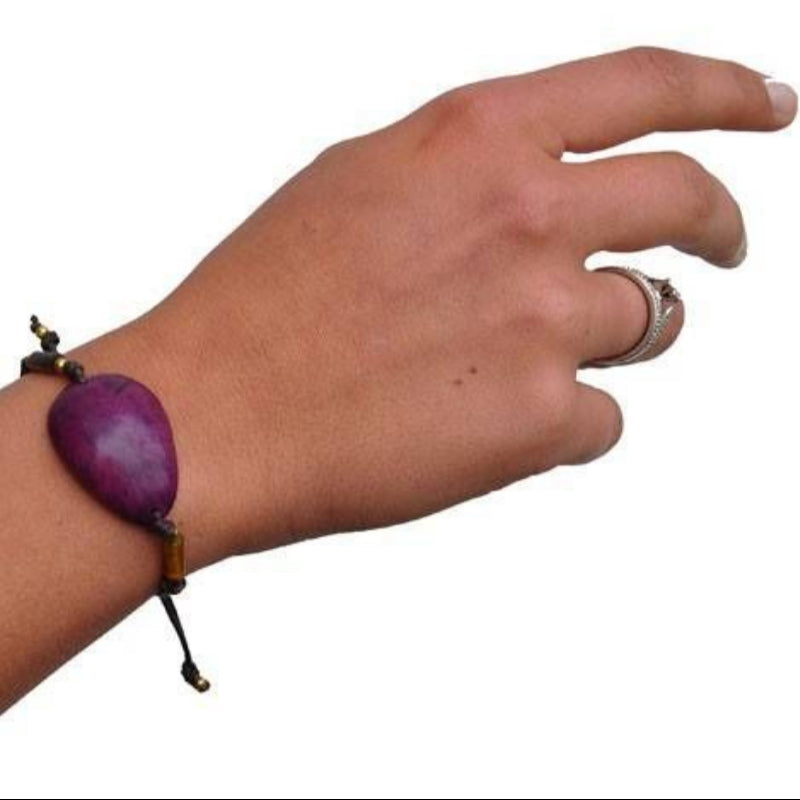 Eine Hand mit hellbrauner Haut, die einen silbernen Ring und ein Armband mit einer großen violetten herzförmigen Perle an einer schwarzen Kordel trägt, vor weißem Hintergrund.