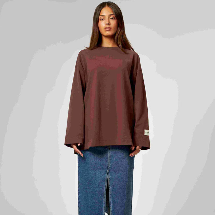 Eine Frau mit langen braunen Haaren trägt ein lockeres, langärmeliges braunes Oberteil und einen blauen Jeansrock mit einem Schlitz an der Vorderseite und steht vor einem schlichten, hellgrauen Hintergrund.