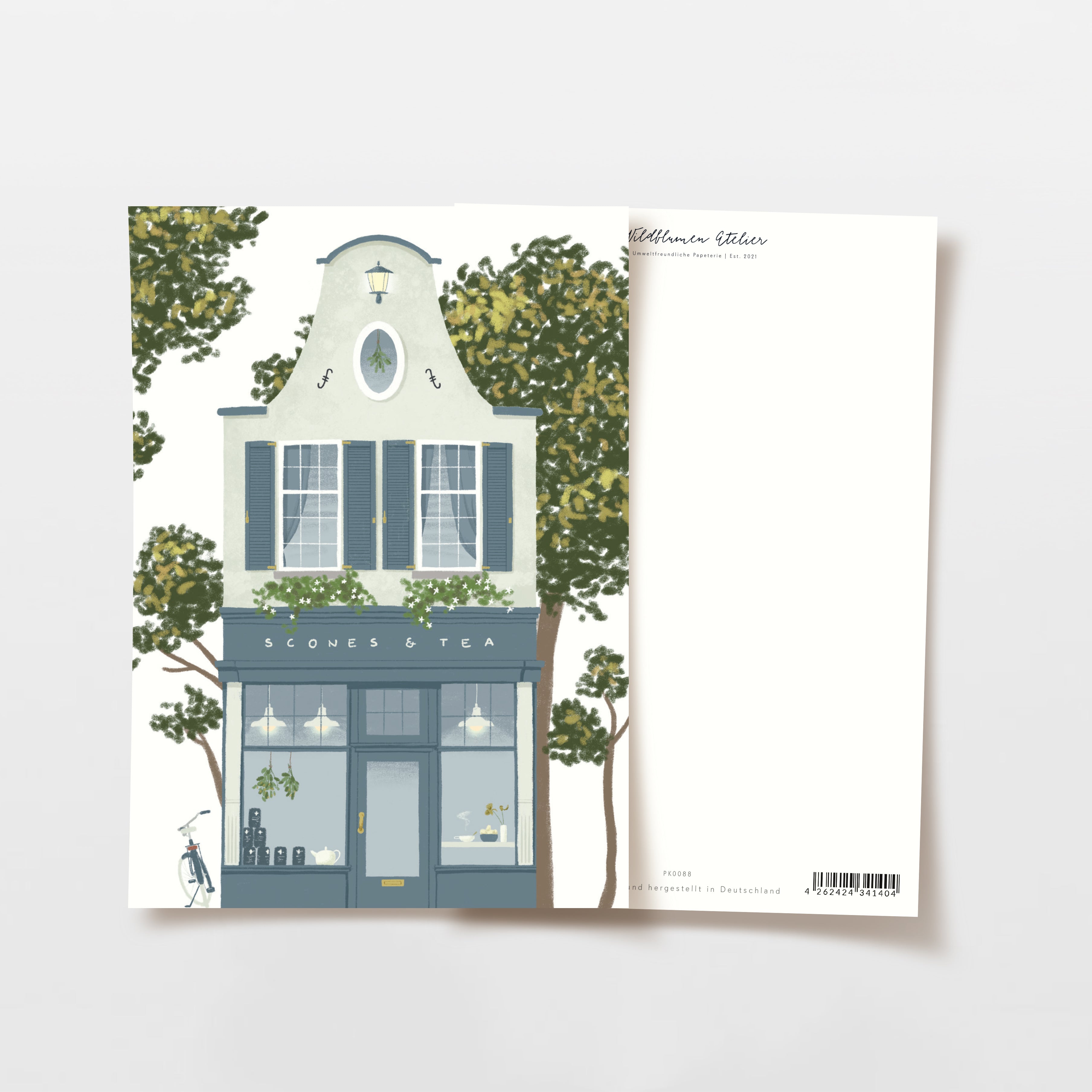 Illustrierte Postkarte, die ein zweistöckiges Geschäft namens Scones & Tea mit Bäumen auf beiden Seiten zeigt. Das Geschäft hat große Fenster, blaugraue Akzente und ein Fahrrad, das draußen geparkt ist. Die Rückseite der Postkarte ist leer, außer für kleine gedruckten Text.