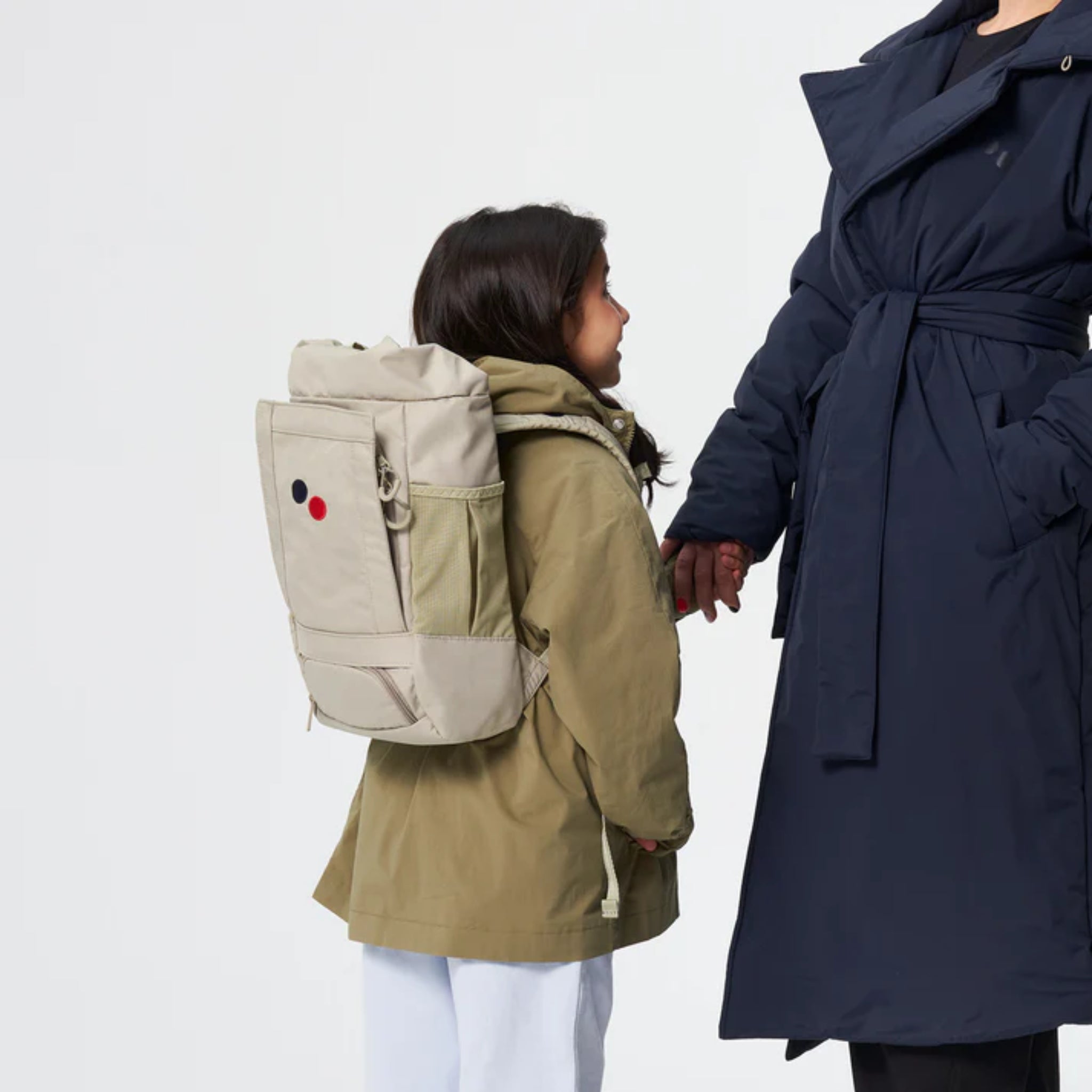 Ein Kind, das eine khakifarbene Jacke und einen beigen Rucksack trägt, hält die Hand eines Erwachsenen in einem marineblauen Mantel. Das Kind schaut zu dem Erwachsenen auf, und der Hintergrund ist einfarbig weiß.