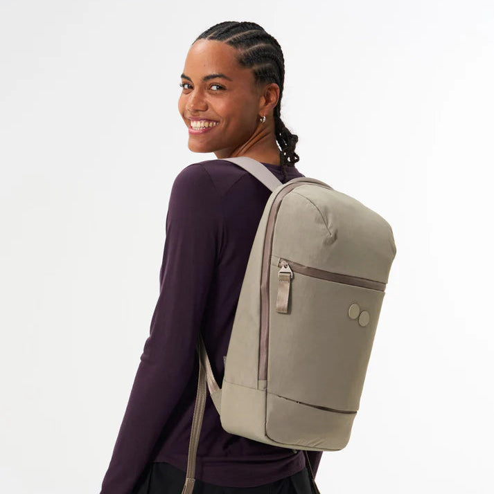 Eine lächelnde Person mit geflochtenen Haaren und einem dunklen langärmeligen Hemd steht seitlich und blickt über die Schulter zurück. Sie trägt einen hellgrauen Rucksack mit zwei kreisförmigen Motiven auf der Vorderseite.