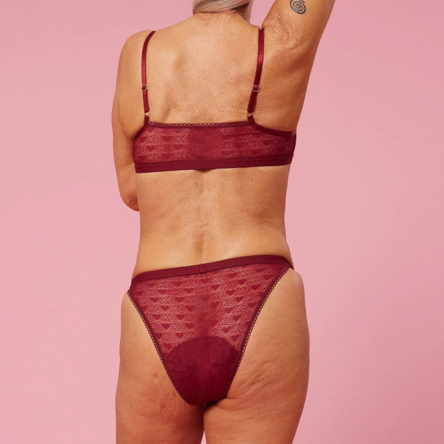 Eine Frau mit grauem Haar, von hinten gesehen, trägt passende kastanienbraune Spitzenunterwäsche vor einem rosa Hintergrund. Sie hat einen Arm erhoben, der eine spiralförmige Tätowierung auf ihrem Oberarm zeigt.