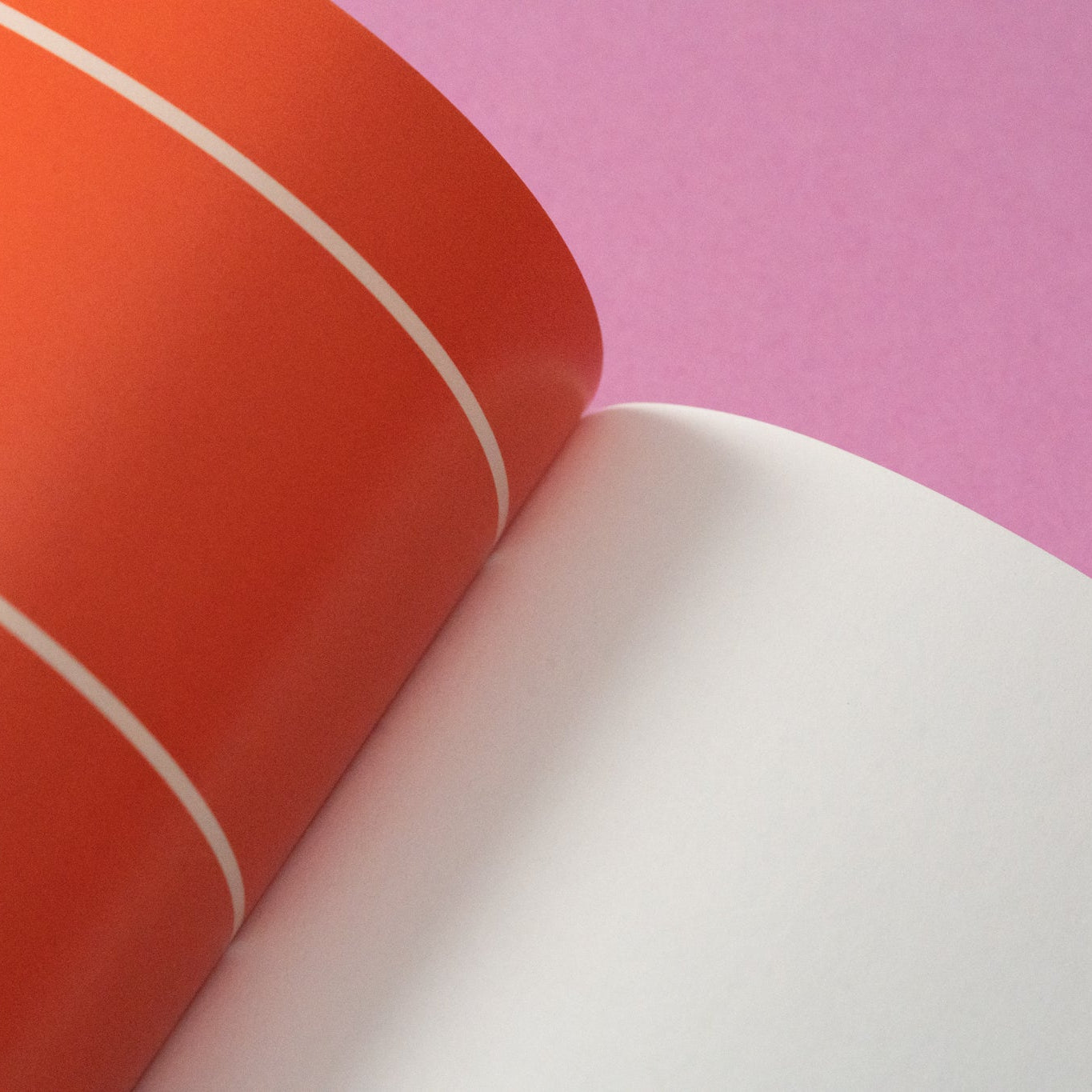 Nahaufnahme eines Buches mit orangefarbenem Einband und zwei weißen Linien, aufgeschlagen auf eine leere weiße Seite, die auf einer rosa Fläche liegt.