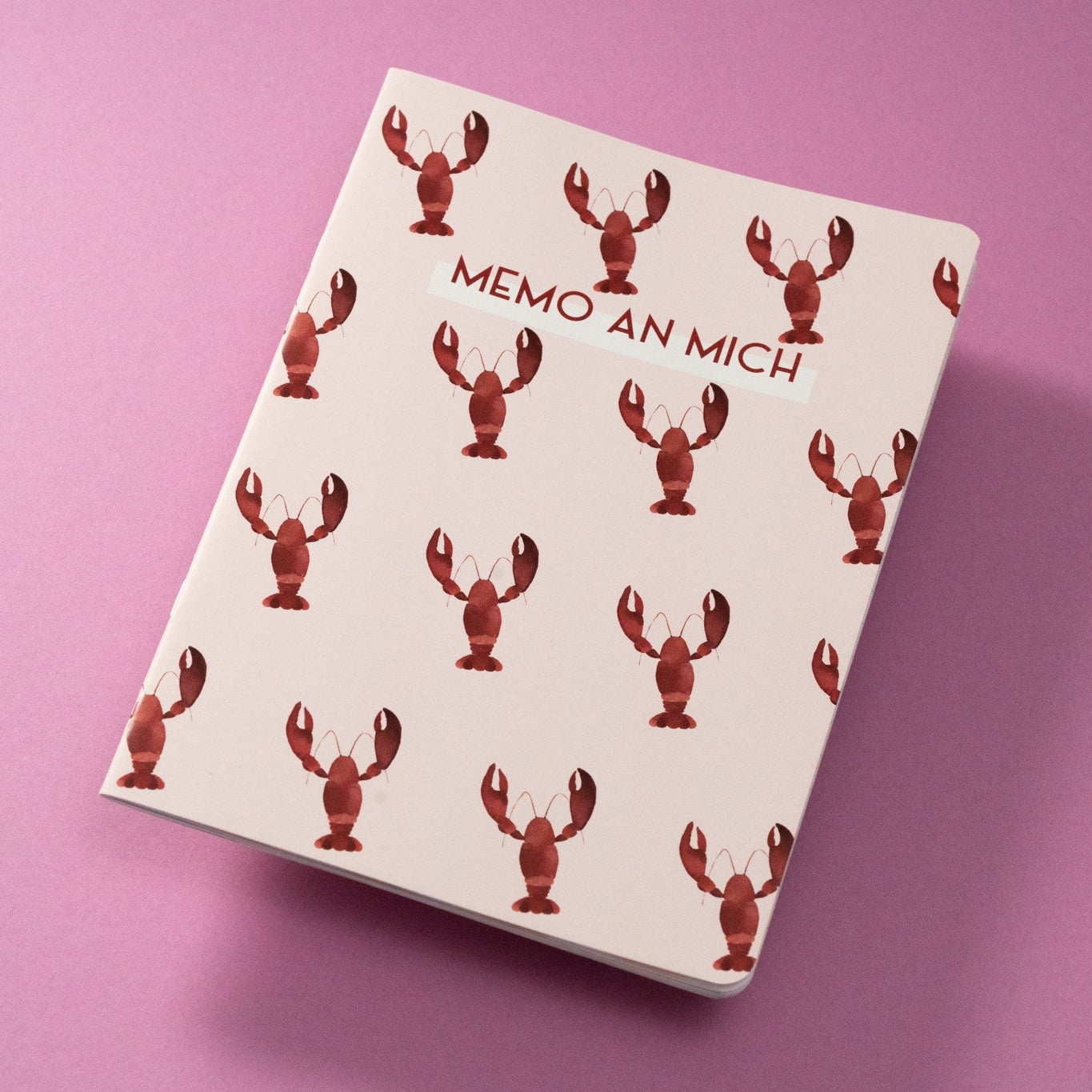 Ein weißes Notizbuch mit roten Hummer-Illustrationen und dem Text MEMO AN MICH auf dem Umschlag, platziert auf einer rosa Oberfläche.