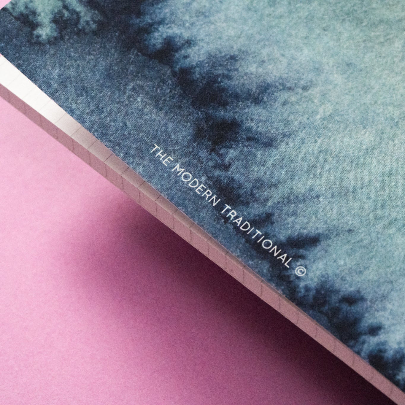 Nahaufnahme eines Buch- oder Notizbucheinbands mit einer blau-grünen Aquarelltextur auf einer rosa Oberfläche. Der Text THE MODERN TRADITIONAL © ist am Rand des Umschlags aufgedruckt.