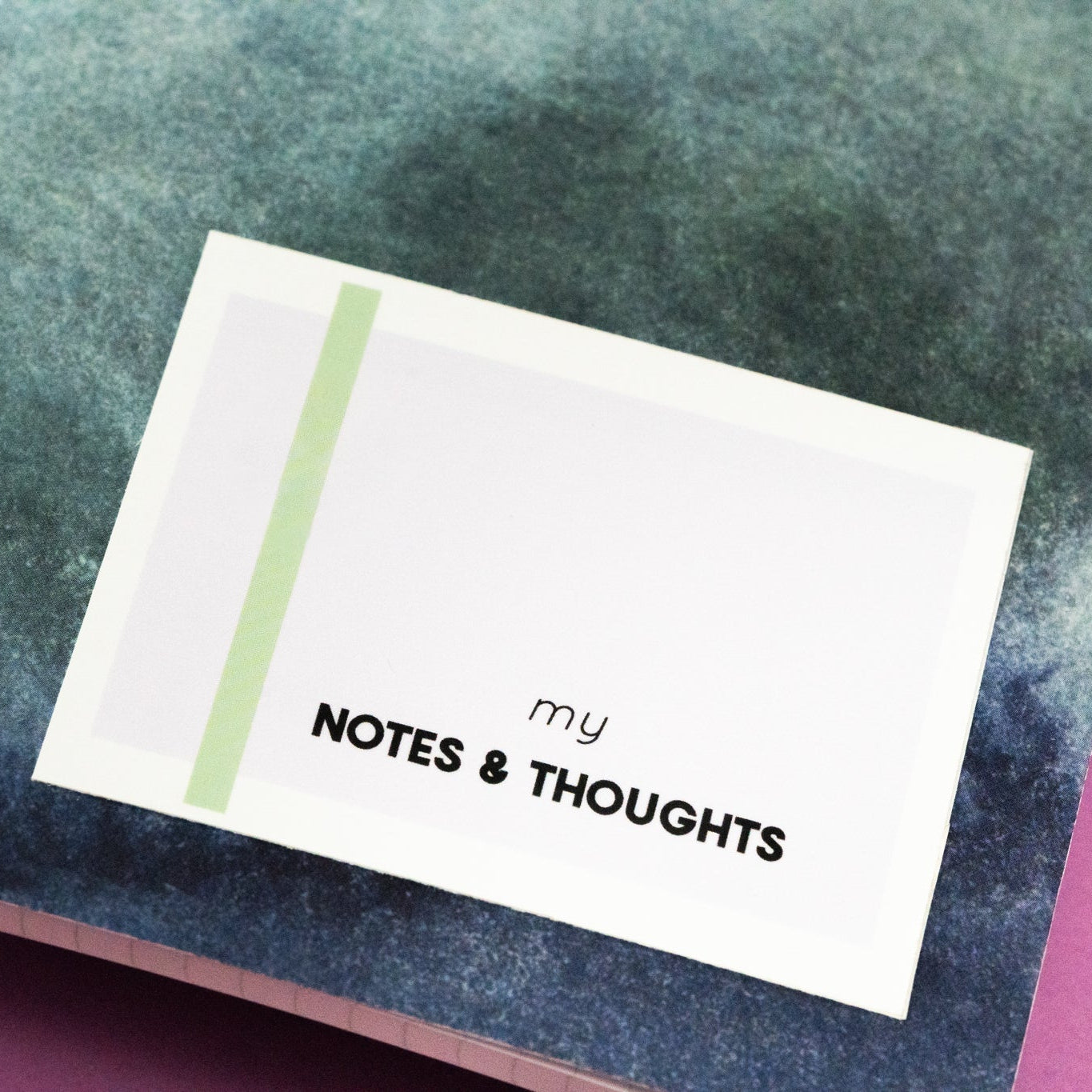 Nahaufnahme eines Notizbuchs mit grünem und blauem Einband im Aquarellstil und einem Etikett mit der Aufschrift MEINE NOTEN & GEDANKEN auf einer rosa Fläche.