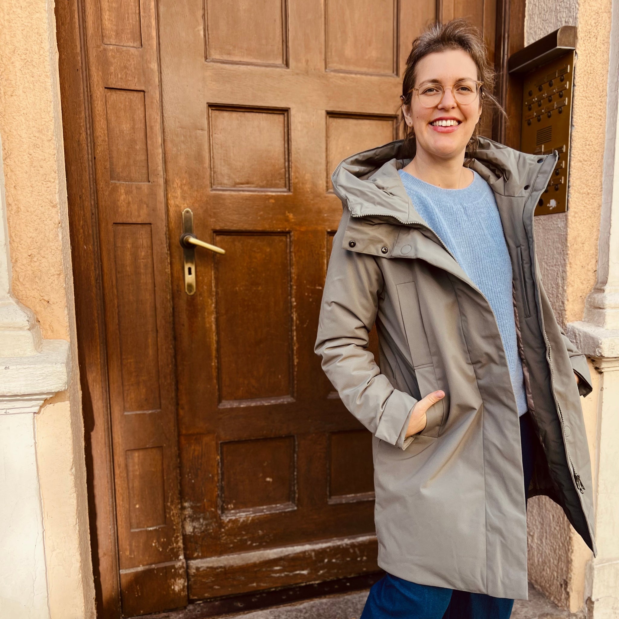Eine Frau in einem grauen Mantel lächelt vor einer Holztür.