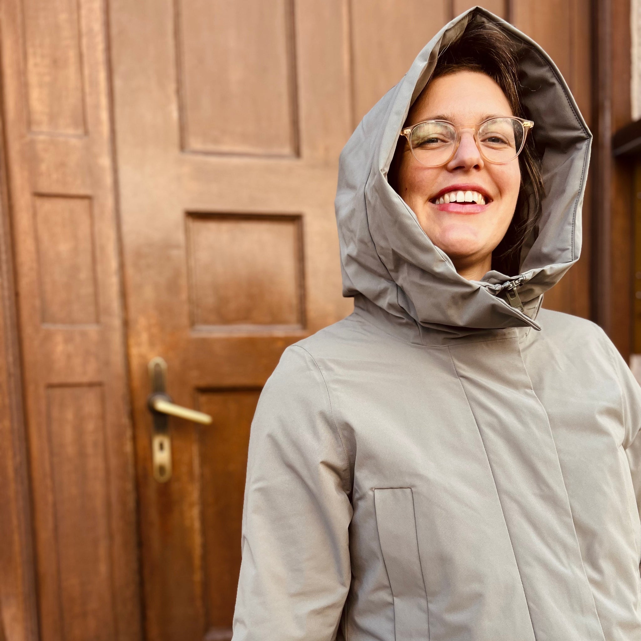 Person mit Brille und grauem Kapuzenmantel lächelt vor einer Holztür.
