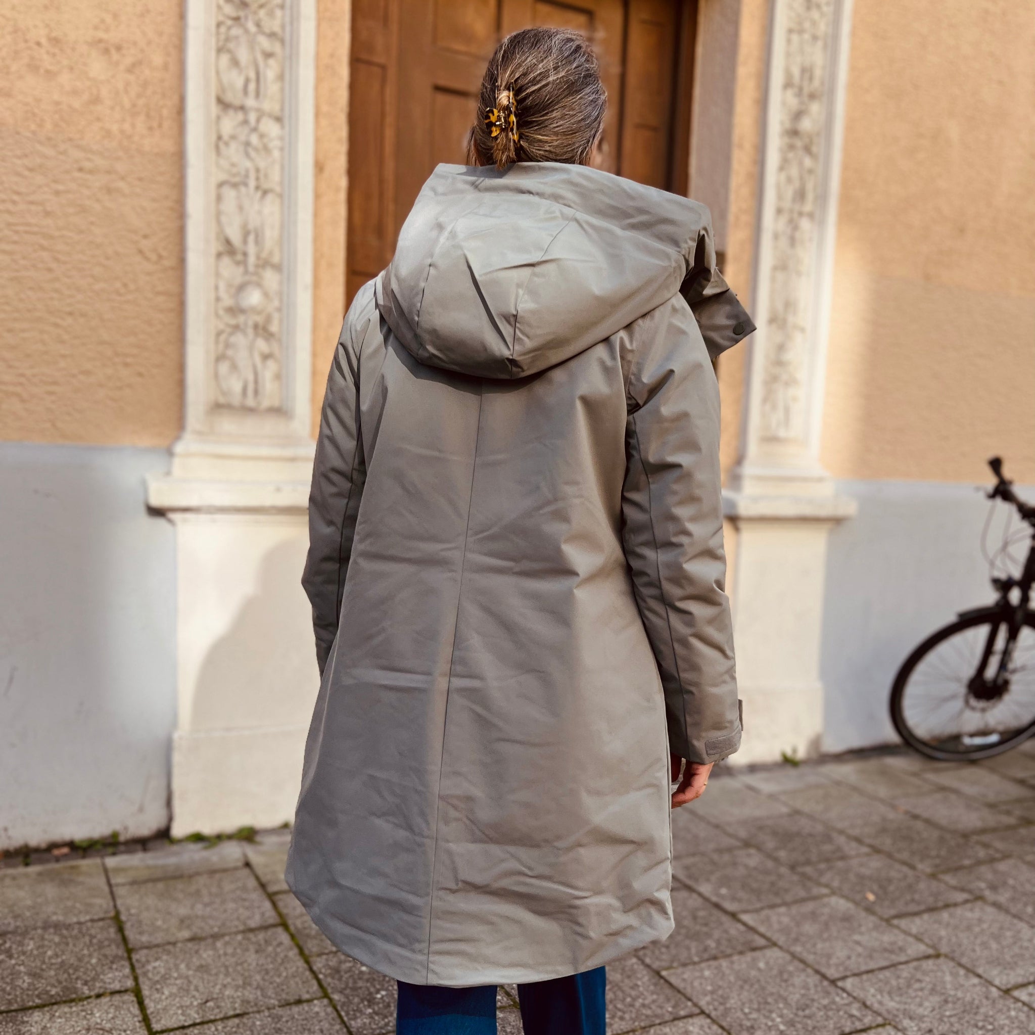 Frau in grauem Mantel steht vor einer verzierten Tür, neben ihr ein Fahrrad.