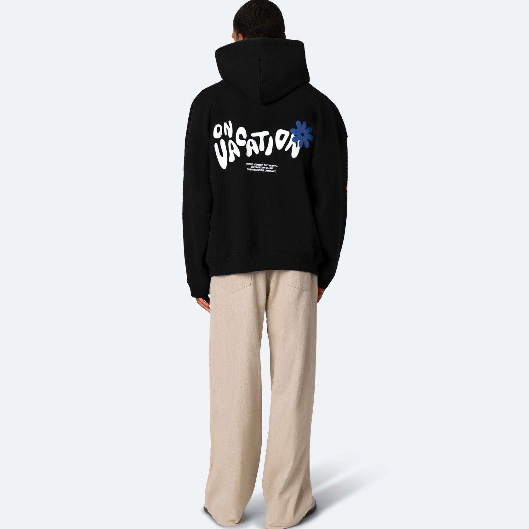 Eine Person steht mit dem Gesicht zur Wand und trägt einen schwarzen Kapuzenpulli mit der Aufschrift ON VACATION und einer blauen Blumengrafik auf dem Rücken, gepaart mit einer lockeren beigen Hose vor einem schlichten hellen Hintergrund.