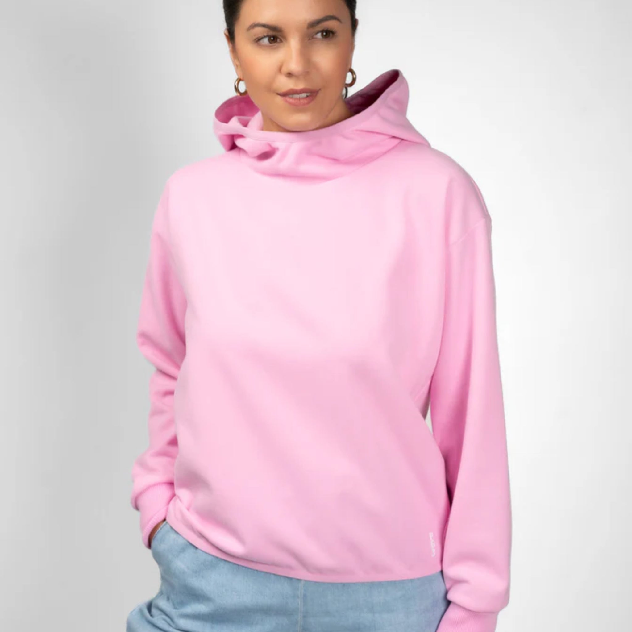 Eine Frau, die ein hellrosa Kapuzensweatshirt und eine hellblaue Hose trägt, steht vor einem schlichten grauen Hintergrund und blickt leicht zur Seite, die Hände in den Taschen.