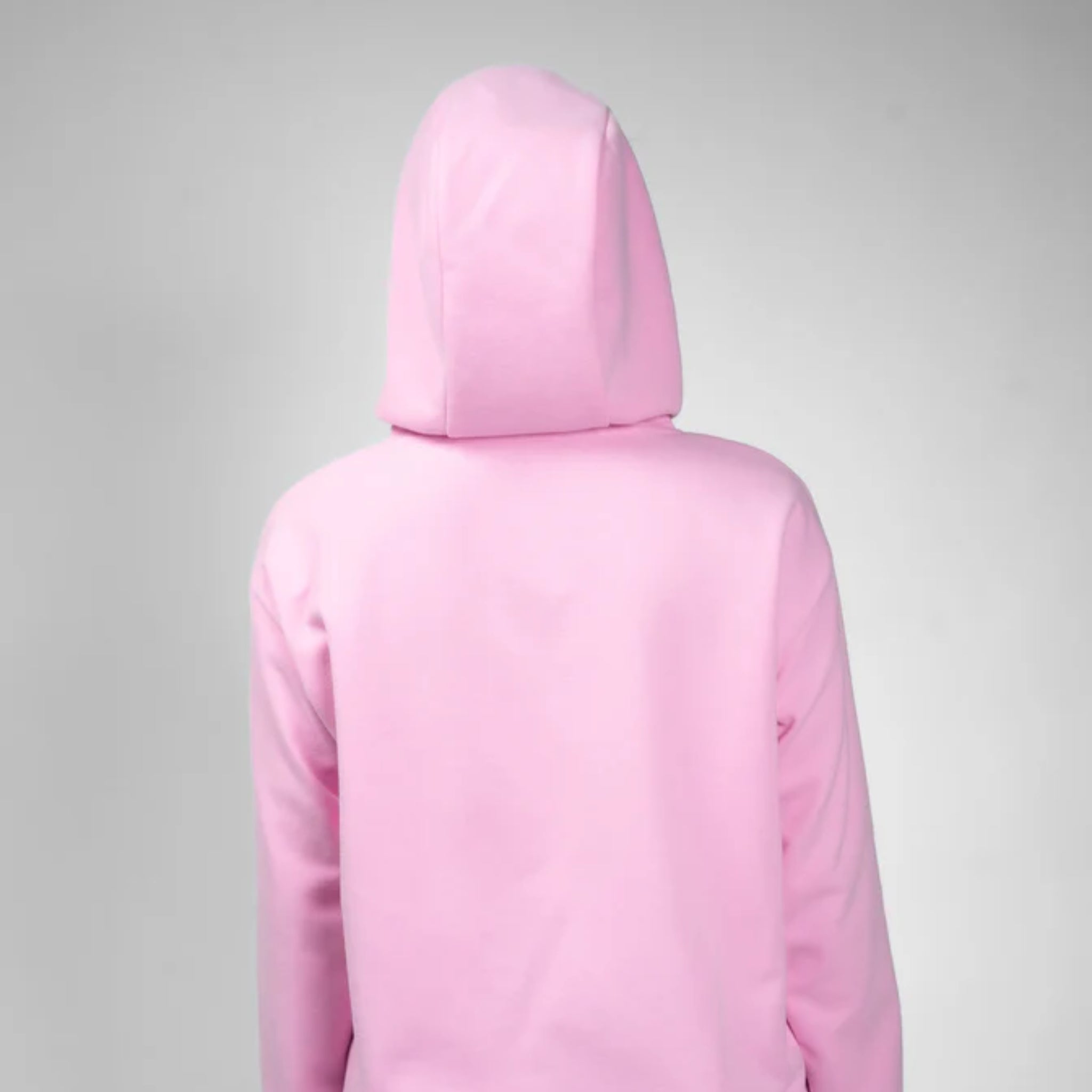 Eine Person in einem hellrosa Kapuzenpulli steht mit dem Gesicht zur Kamera vor einem schlichten grauen Hintergrund.