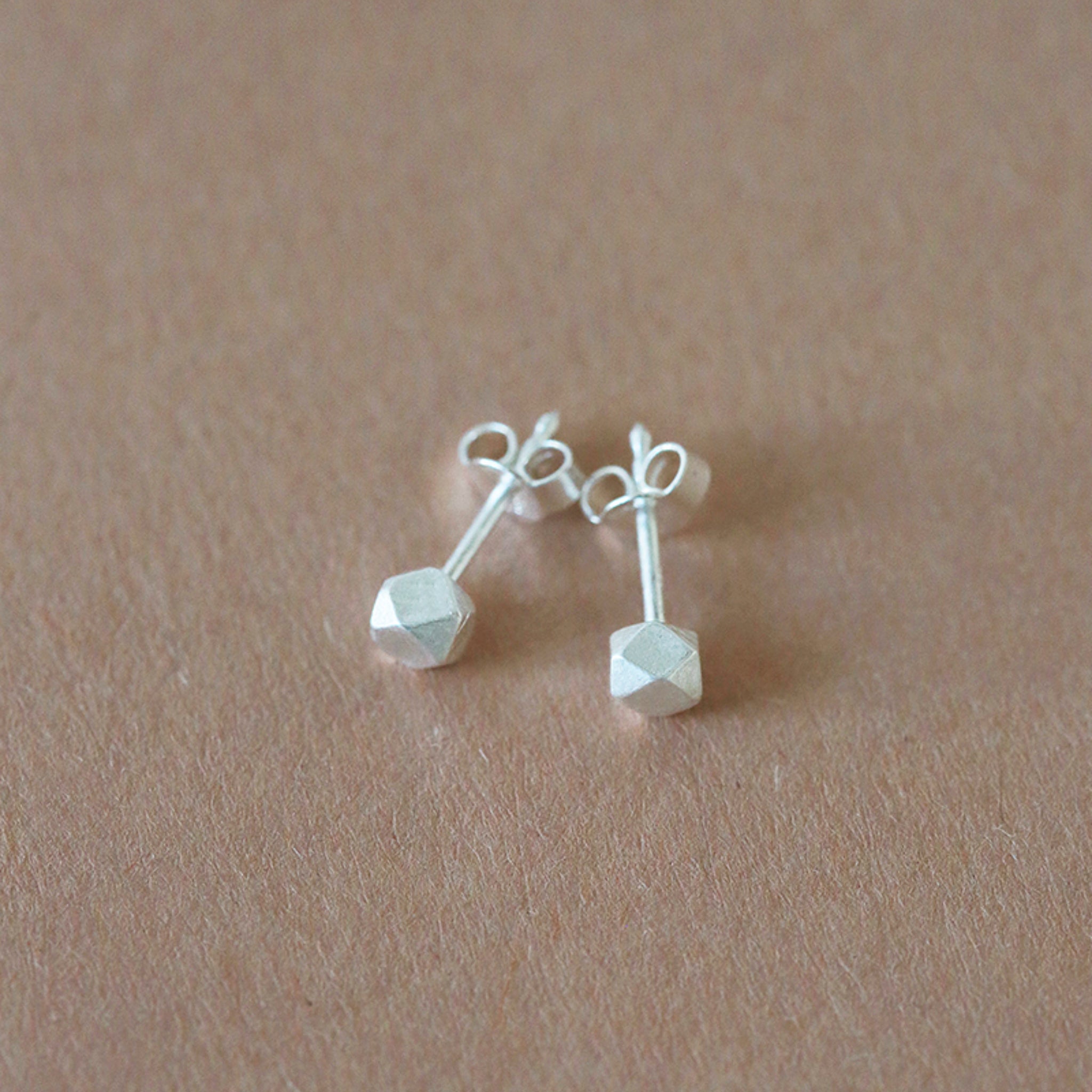Ein Paar kleiner, geometrischer Silberohrstecker mit sechseckigen Enden, die auf einer hellbraunen, strukturierten Oberfläche liegen.