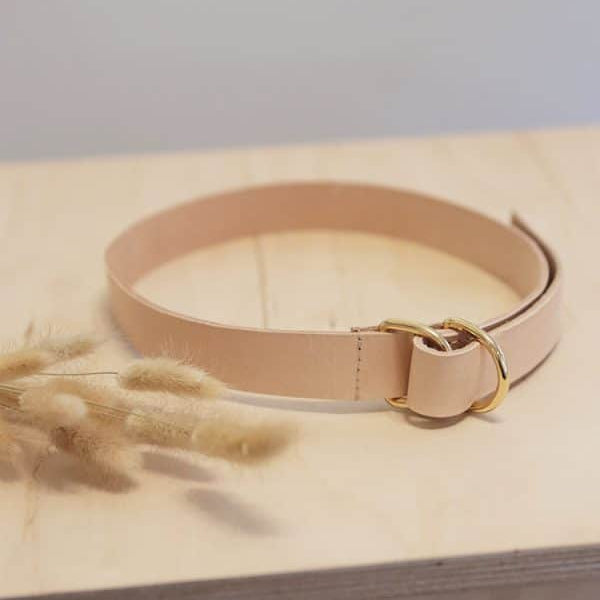 Ein hellbrauner Ledergürtel mit einer goldenen Schnalle liegt auf einer hellen Holzfläche neben einem kleinen Bündel getrockneter beiger Gräser.