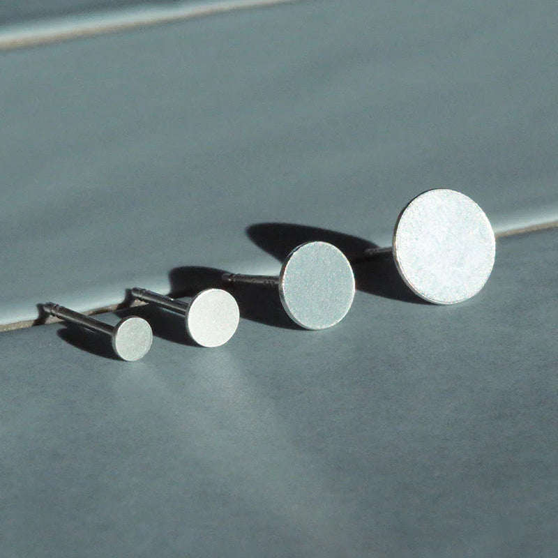 Drei silberne Ohrstecker in ansteigender Größe sind in einer Reihe auf einer glatten, hellgrauen Oberfläche angeordnet und werfen weiche Schatten. Die Ohrringe sind einfach, rund und minimalistisch gestaltet.